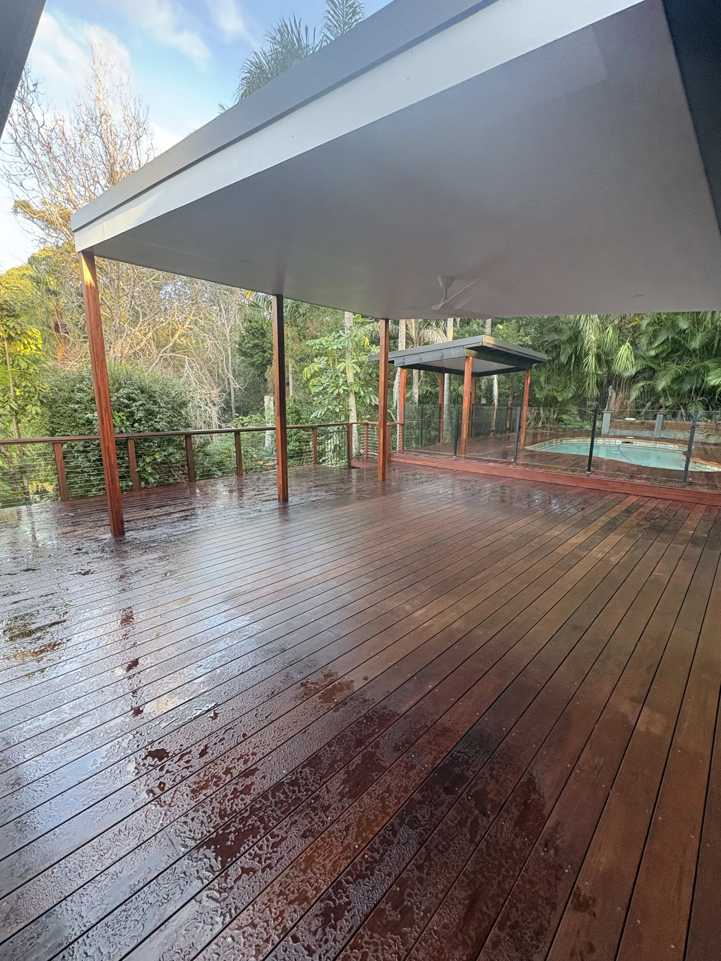 Wet wooden deck with rain reflections, covered patio area, trees and cloudy sky in the background.