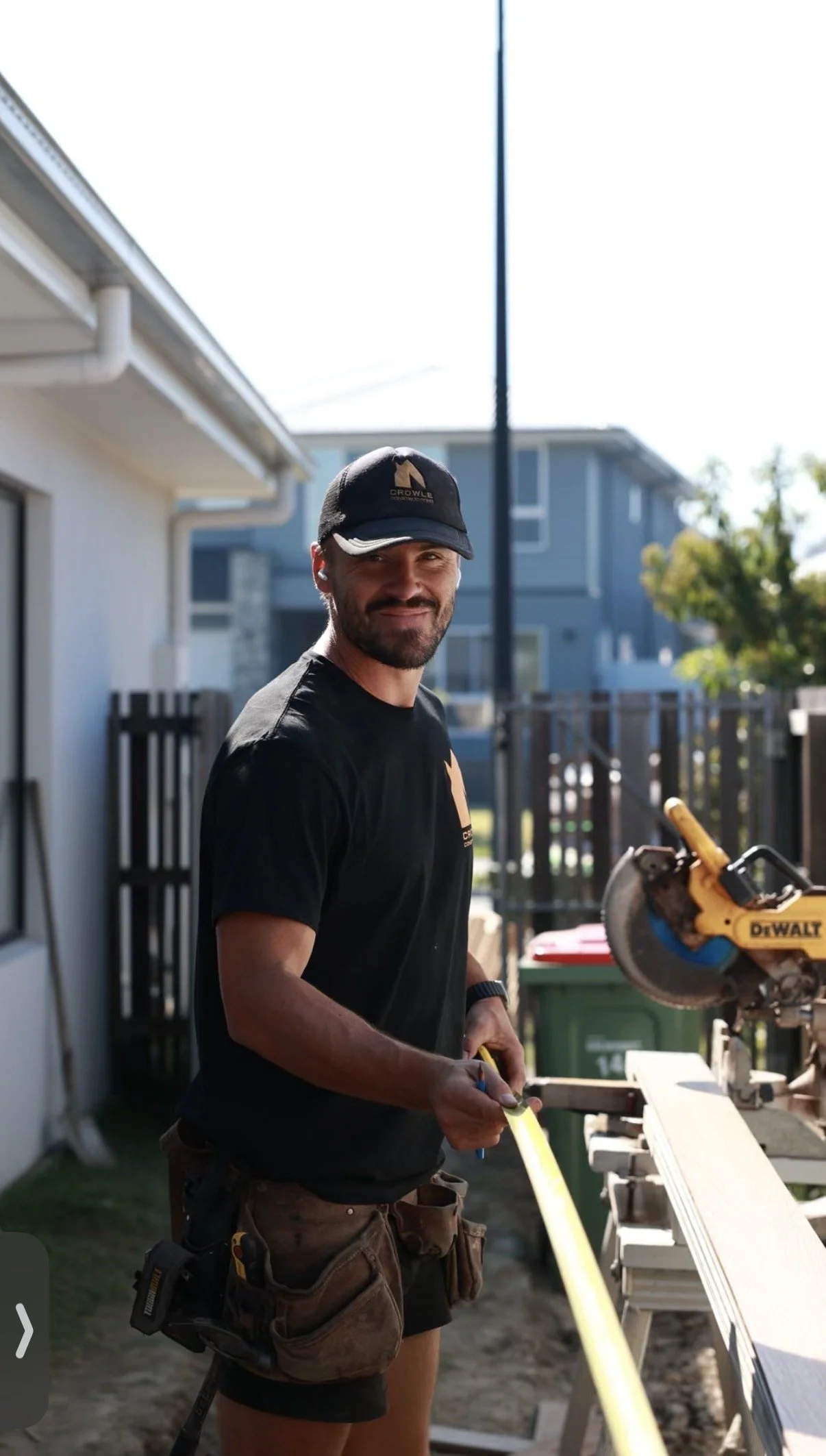 A man with a beard and a black cap is working outdoors in a residential backyard, measuring a wooden board with a tape measure. He is wearing a black t-shirt and brown work shorts with tools attached to his belt. There is a saw on the work table next to him, and houses and trees are visible in the background.
