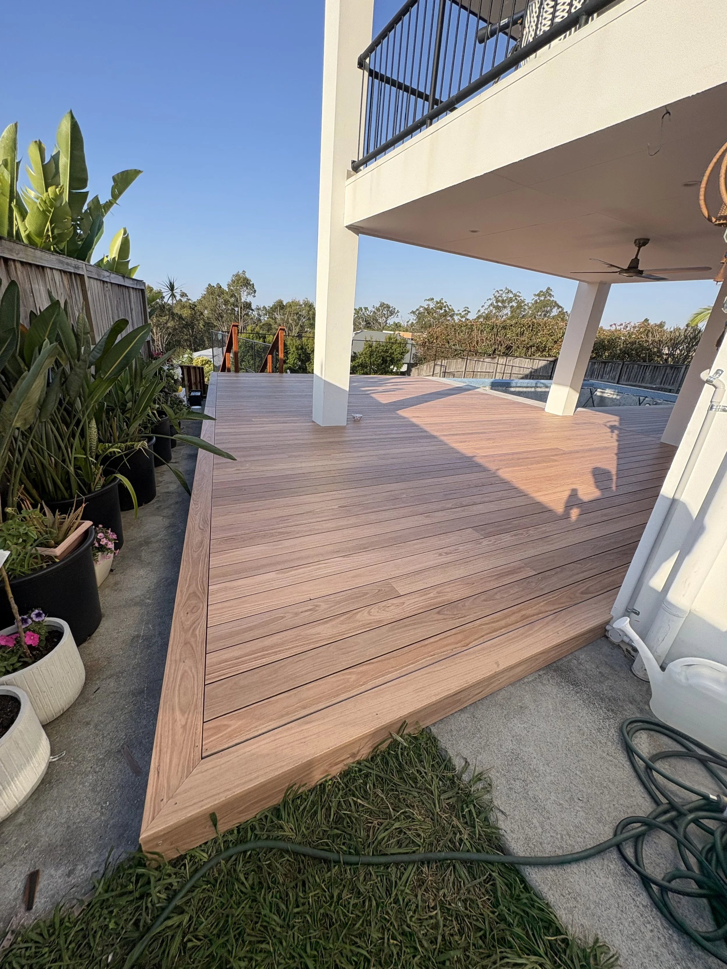 Backyard deck with wooden flooring, potted plants on the left, house with balcony on the right, outdoor ceiling fan, fenced backyard, trees, and clear blue sky.
