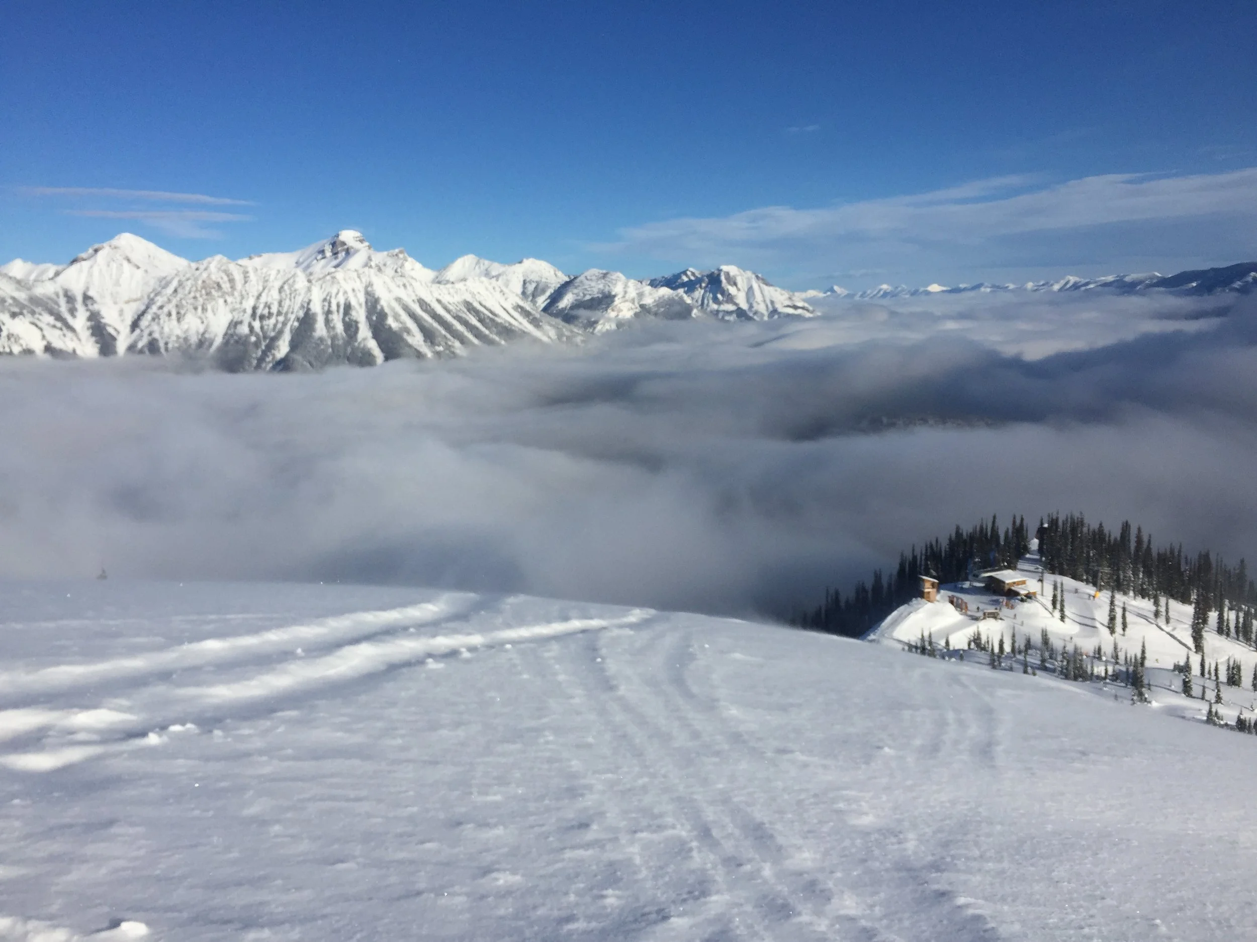 Snow-covered mountains with a blue sky and clouds, and a small building surrounded by trees in the foreground.