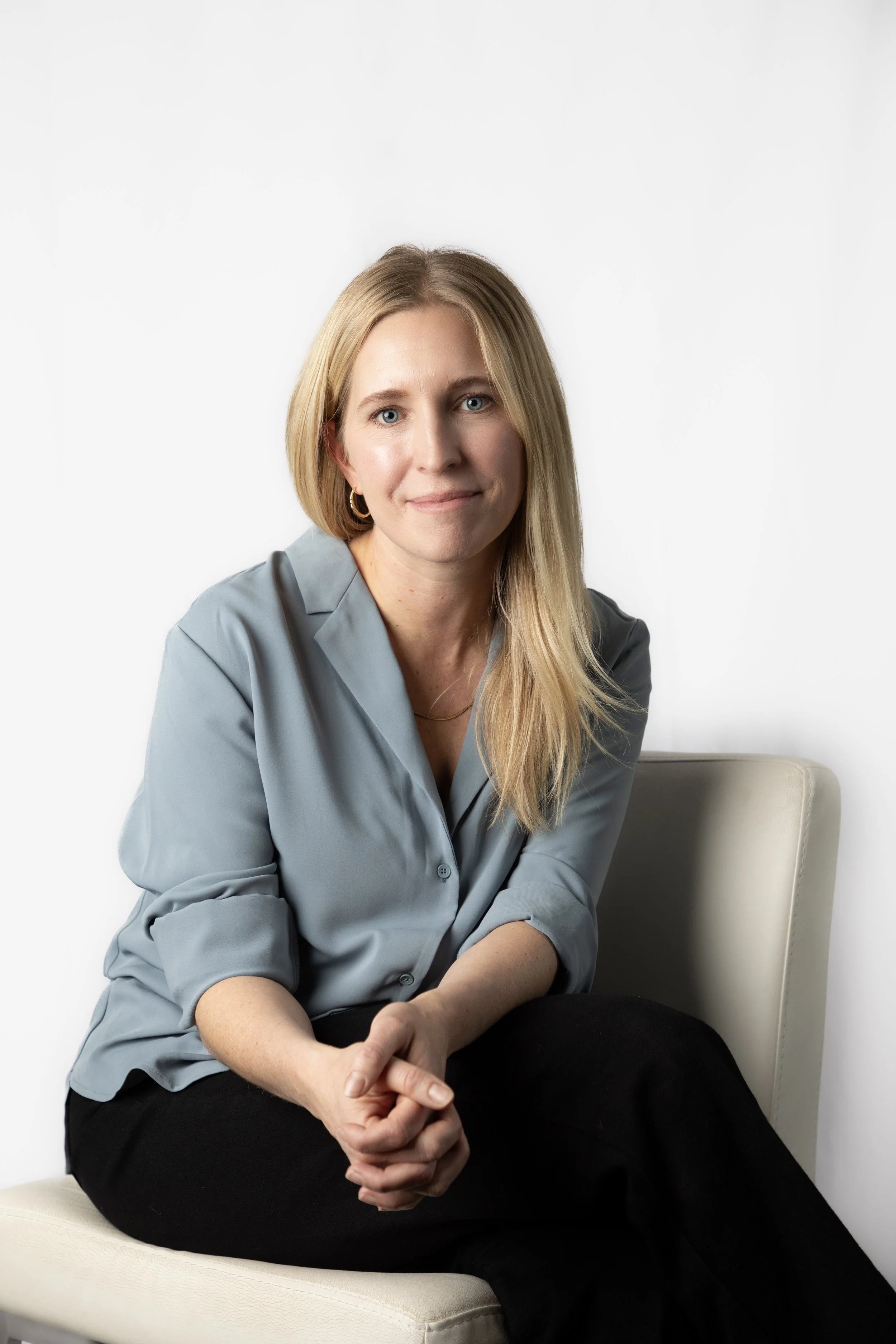 A woman with blonde hair, wearing a gray button-up shirt, sitting on a cream-colored chair against a plain white background, with her hands clasped in her lap.