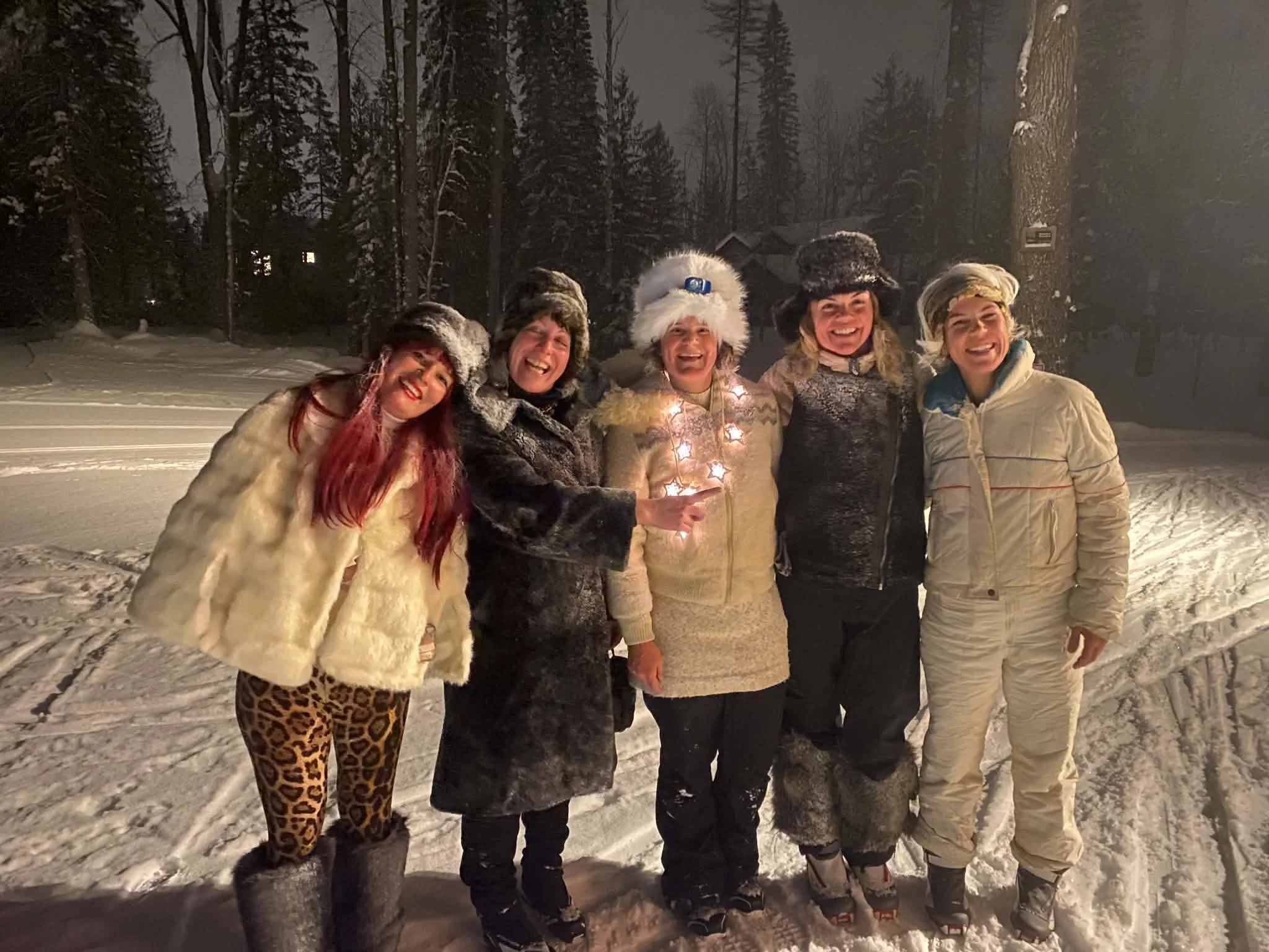 A group of five women standing close together outdoors at night in winter, dressed in warm clothing and wearing festive hats, smiling at the camera, with snow-covered ground and tall trees in the background.