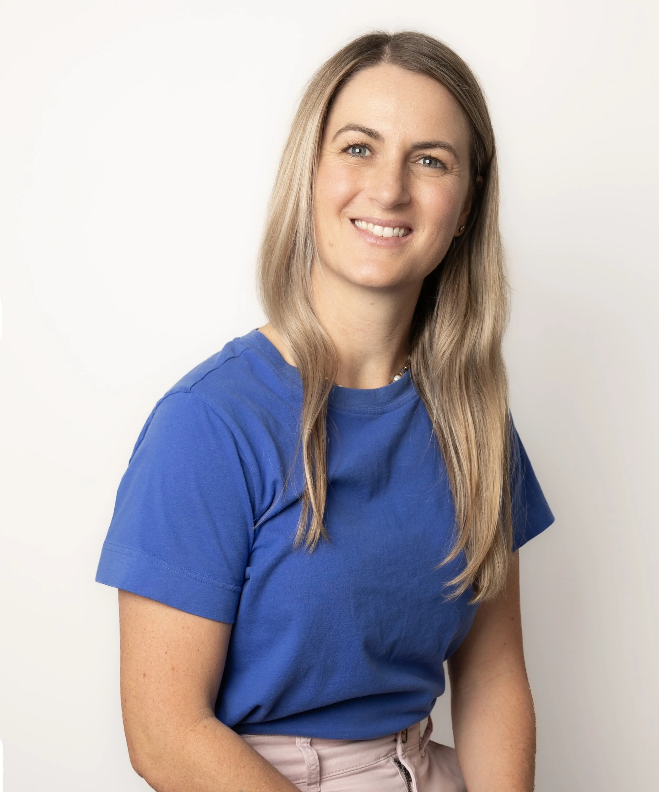 A woman with long blonde hair wearing a blue T-shirt and light-colored pants, smiling against a plain white background.