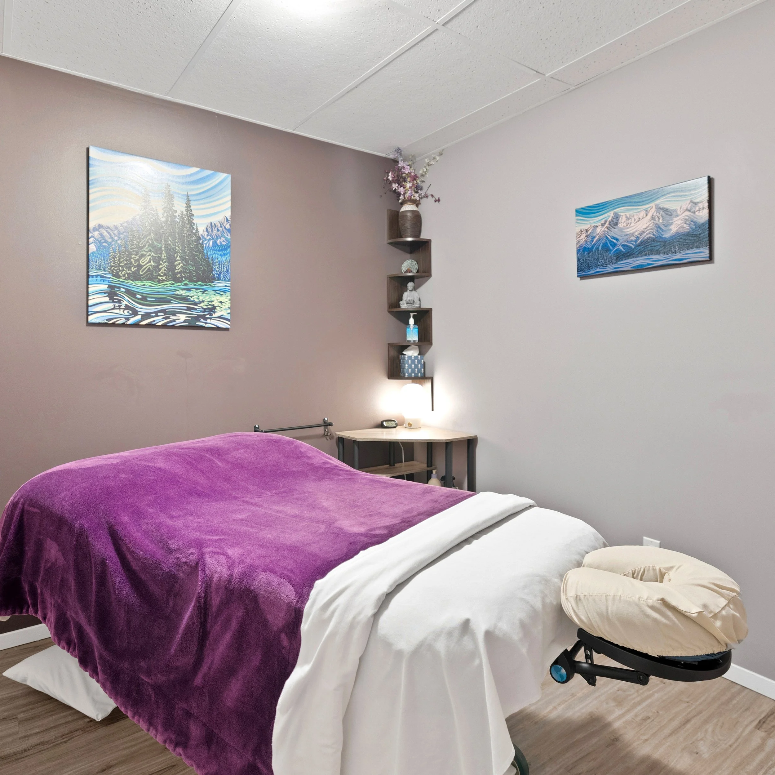 A massage room with a massage bed draped with a purple blanket and a white sheet, a massage pillow, a small bedside table with a clock and a soap dispenser, framed mountain and lake landscape paintings on the wall, a corner shelf with floral arrangement, a small Buddha statue, and a bottle of sanitizer or lotion.