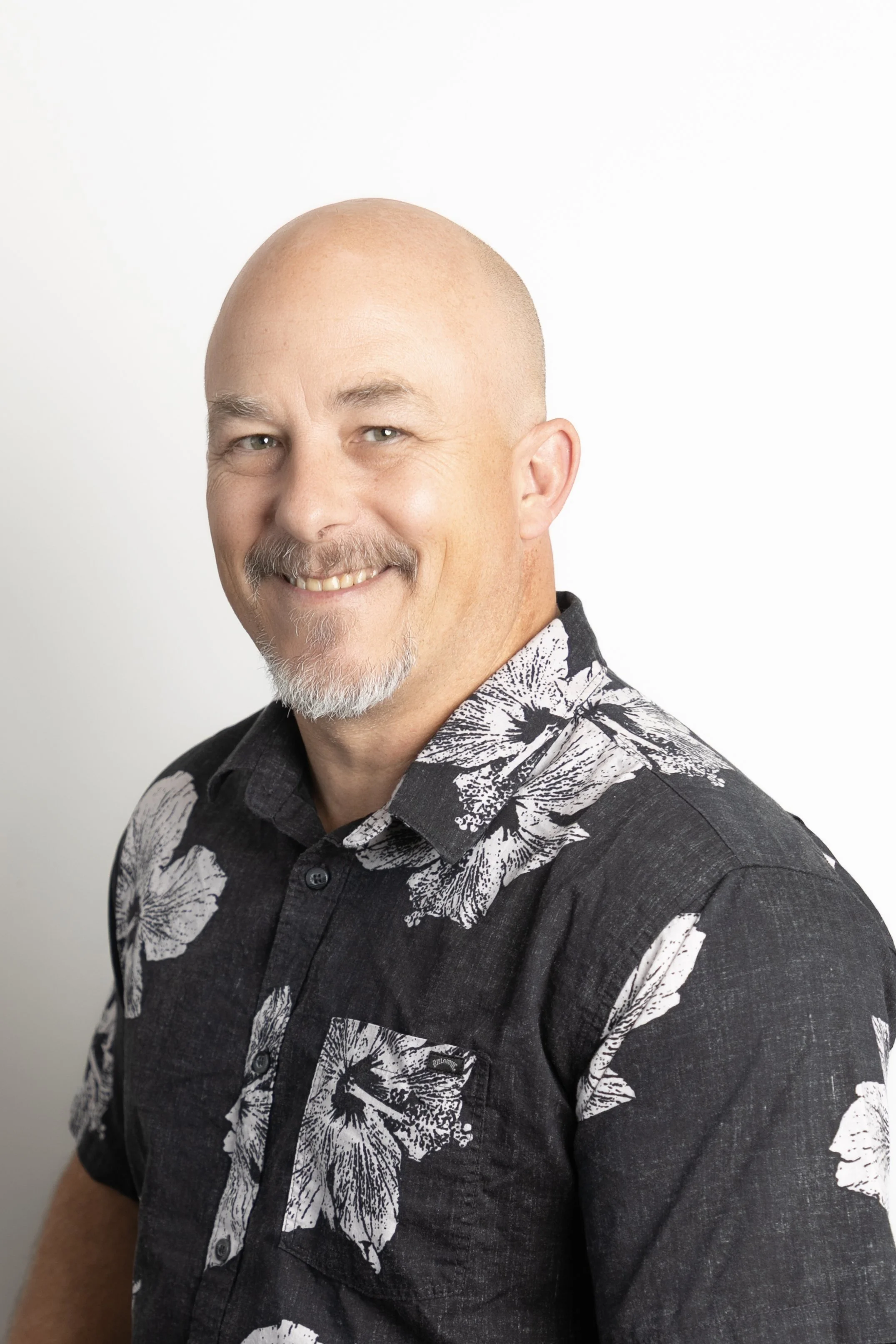 A smiling bald man with a goatee and mustache wearing a black short sleeve shirt with a white floral pattern, standing against a plain white background.