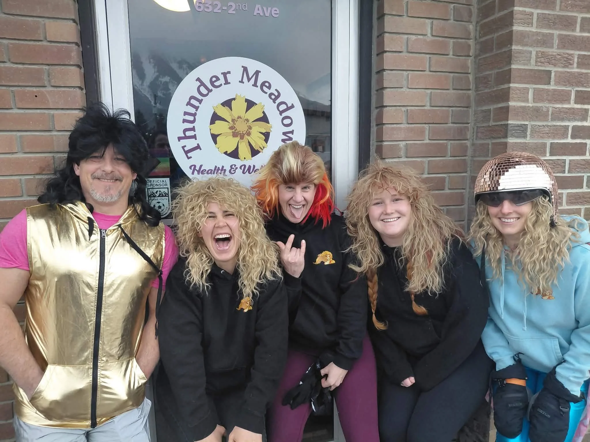 Group of people dressed in colorful costumes, including one with a gold vest, another with a red and orange wig, and another with a gold disco ball hat, standing in front of a building with a sign that reads 'Thunderdome Health & Wellness'.