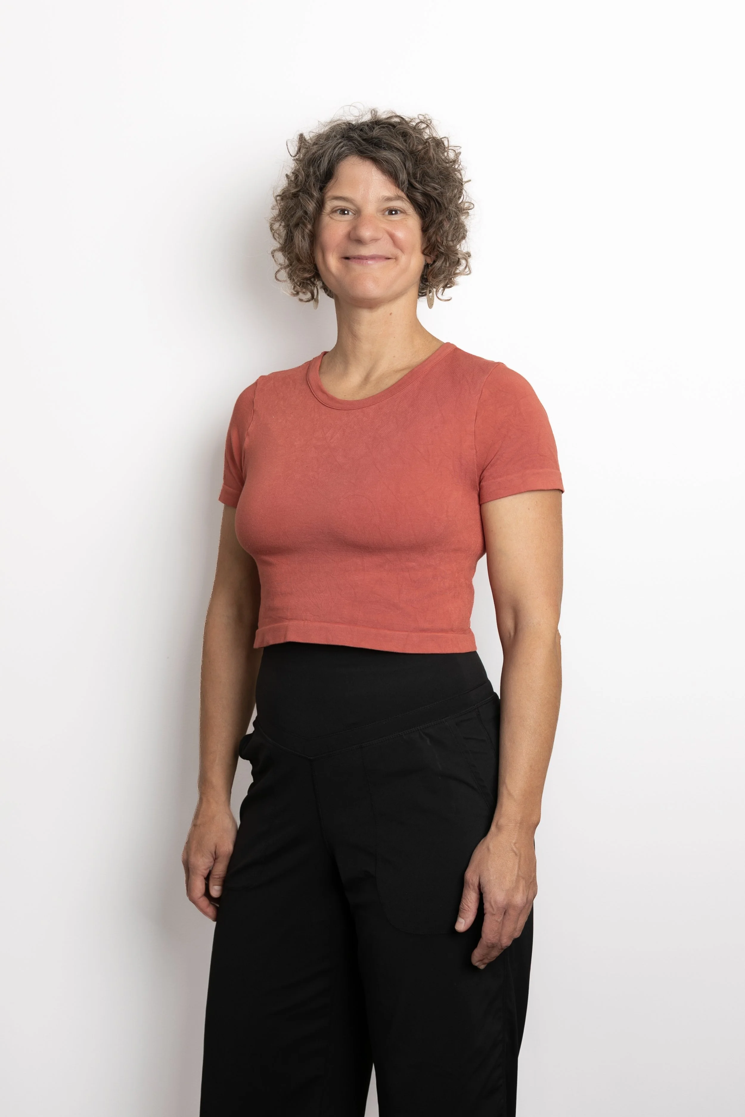 A woman with curly hair, wearing a reddish-orange short-sleeved top and black pants, standing against a plain white background.