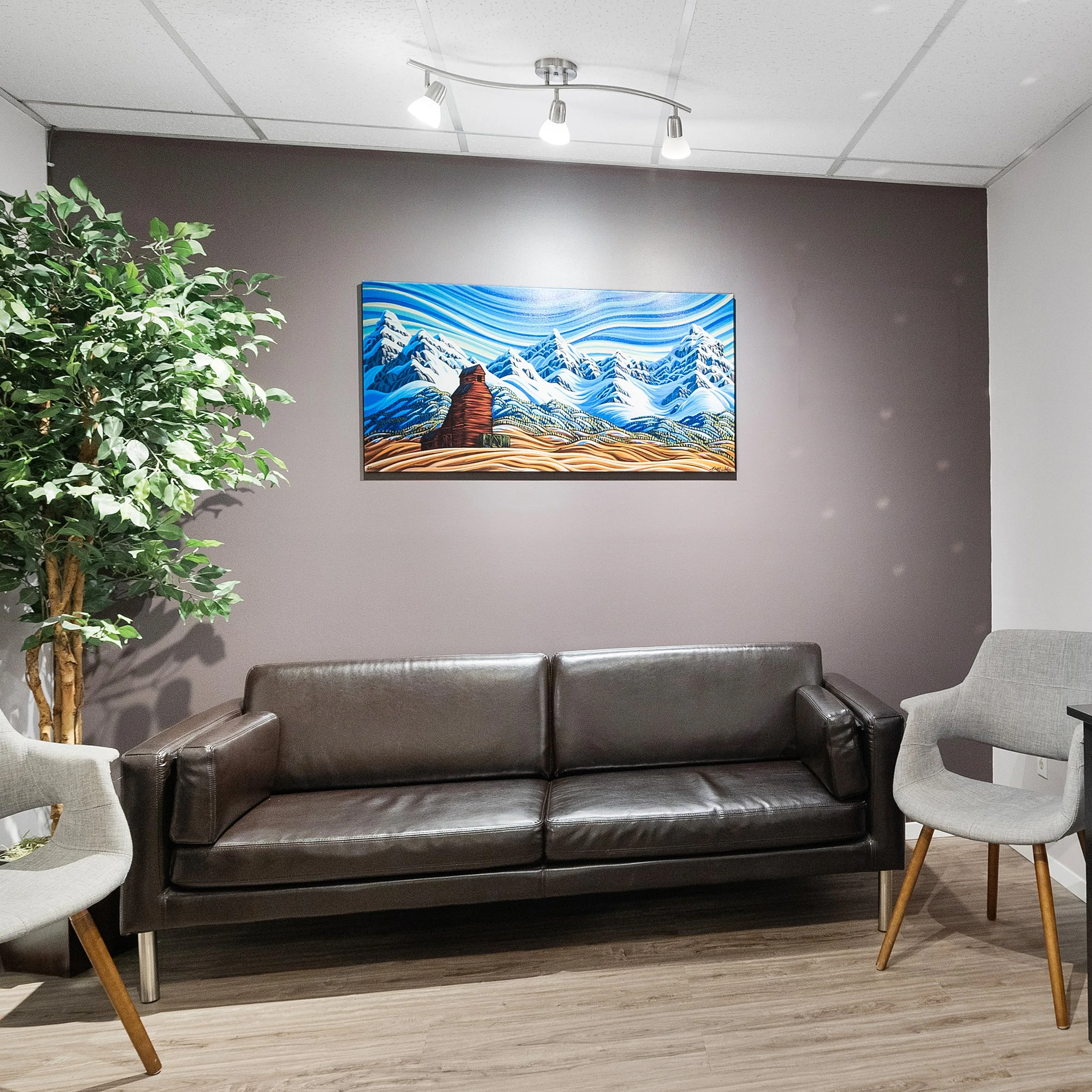 A waiting room with a black sofa, a white armchair, a gray armchair, a wooden chair, a large green leafy plant, a colorful painting of mountains, snow, and a wooden pathway, and ceiling lights.