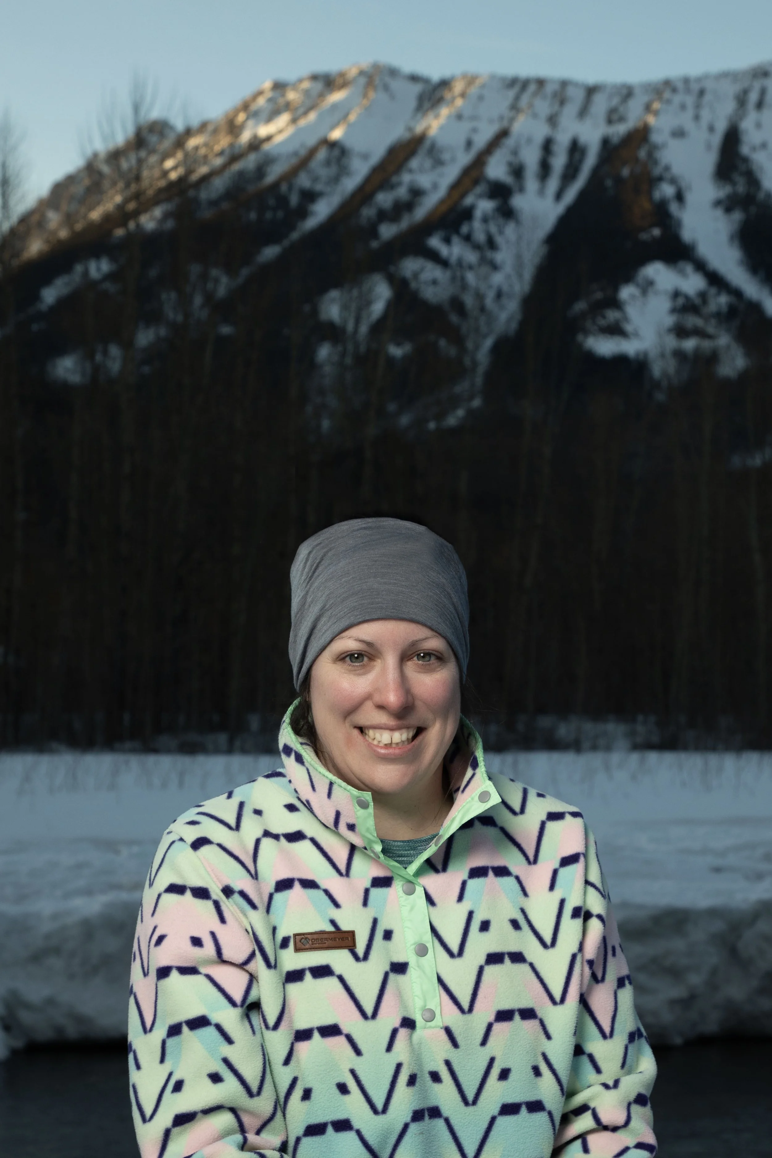 A smiling woman wearing a gray headband and a patterned jacket standing outdoors with snow-covered mountains and trees in the background.