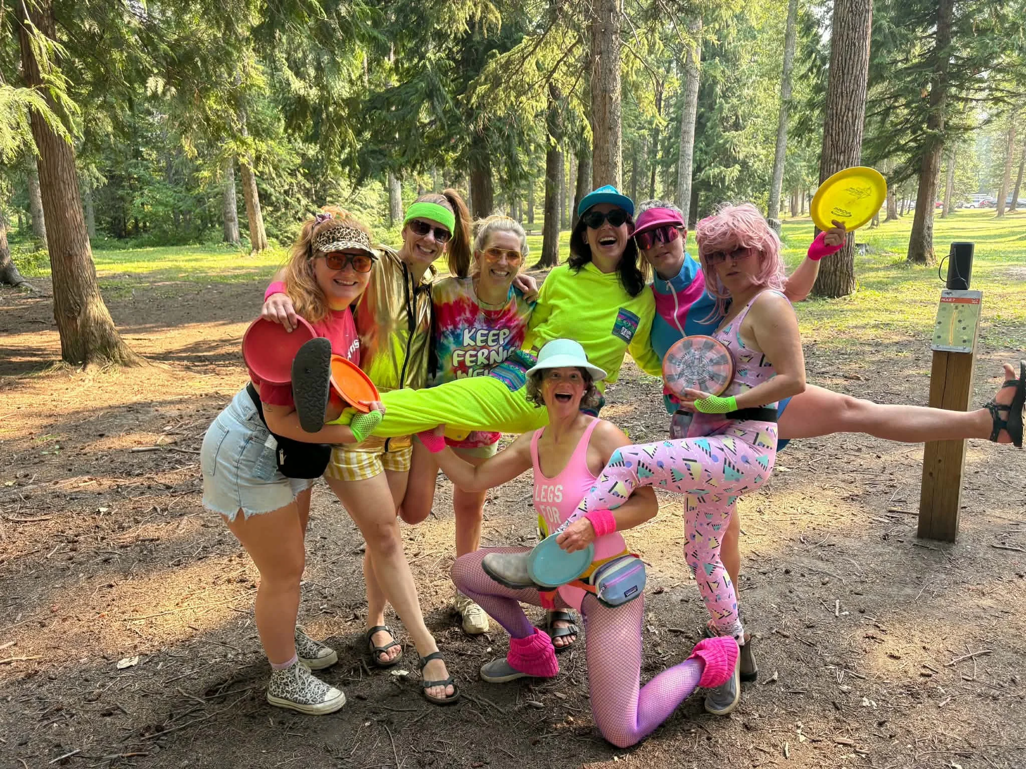 A group of women dressed in colorful, eccentric clothing and accessories are smiling and posing together in a wooded park, holding frisbees and other outdoor gear.