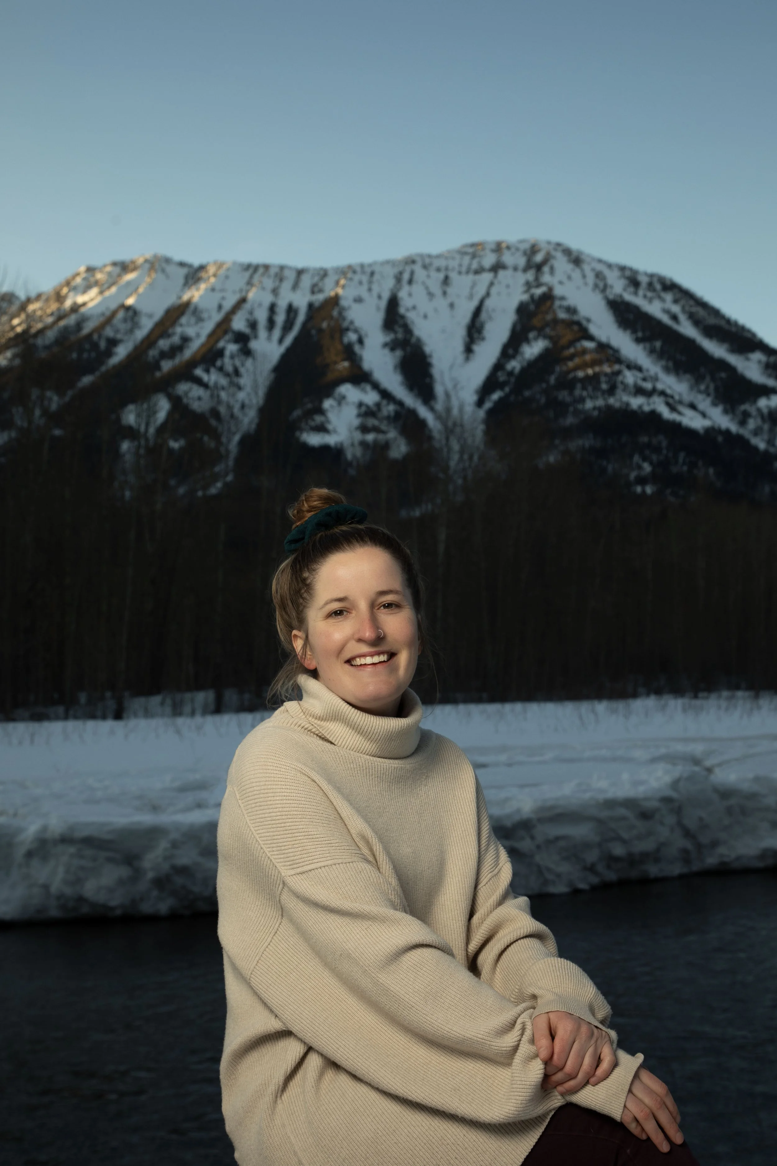 A young woman with light brown hair tied up in a bun, wearing a beige turtleneck sweater, sitting by a river with snow on its banks. Snow-capped mountains are visible in the background under a clear sky.