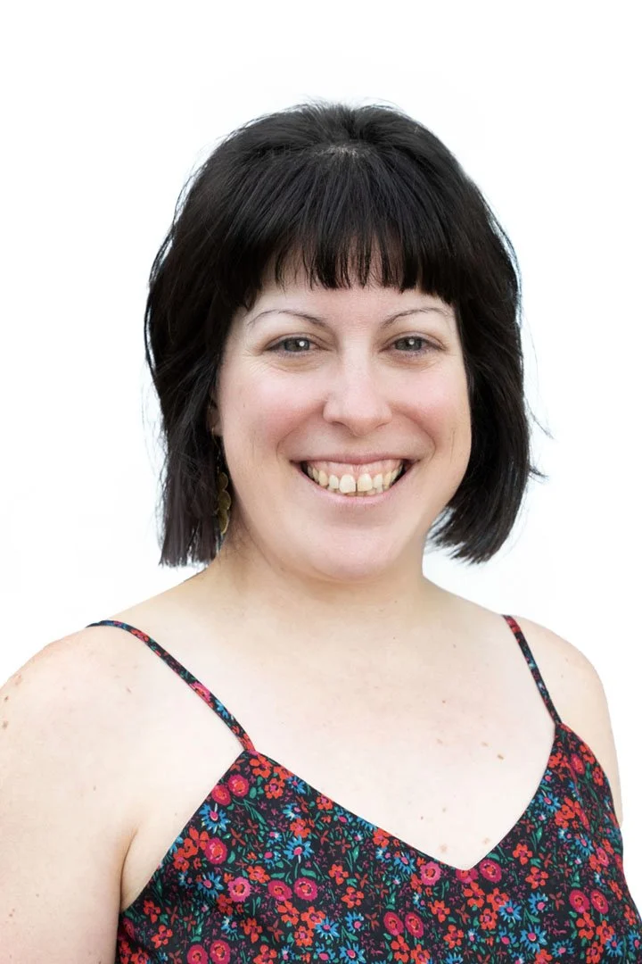 A woman with short dark hair smiling, wearing a colorful floral dress with spaghetti straps against a white background.