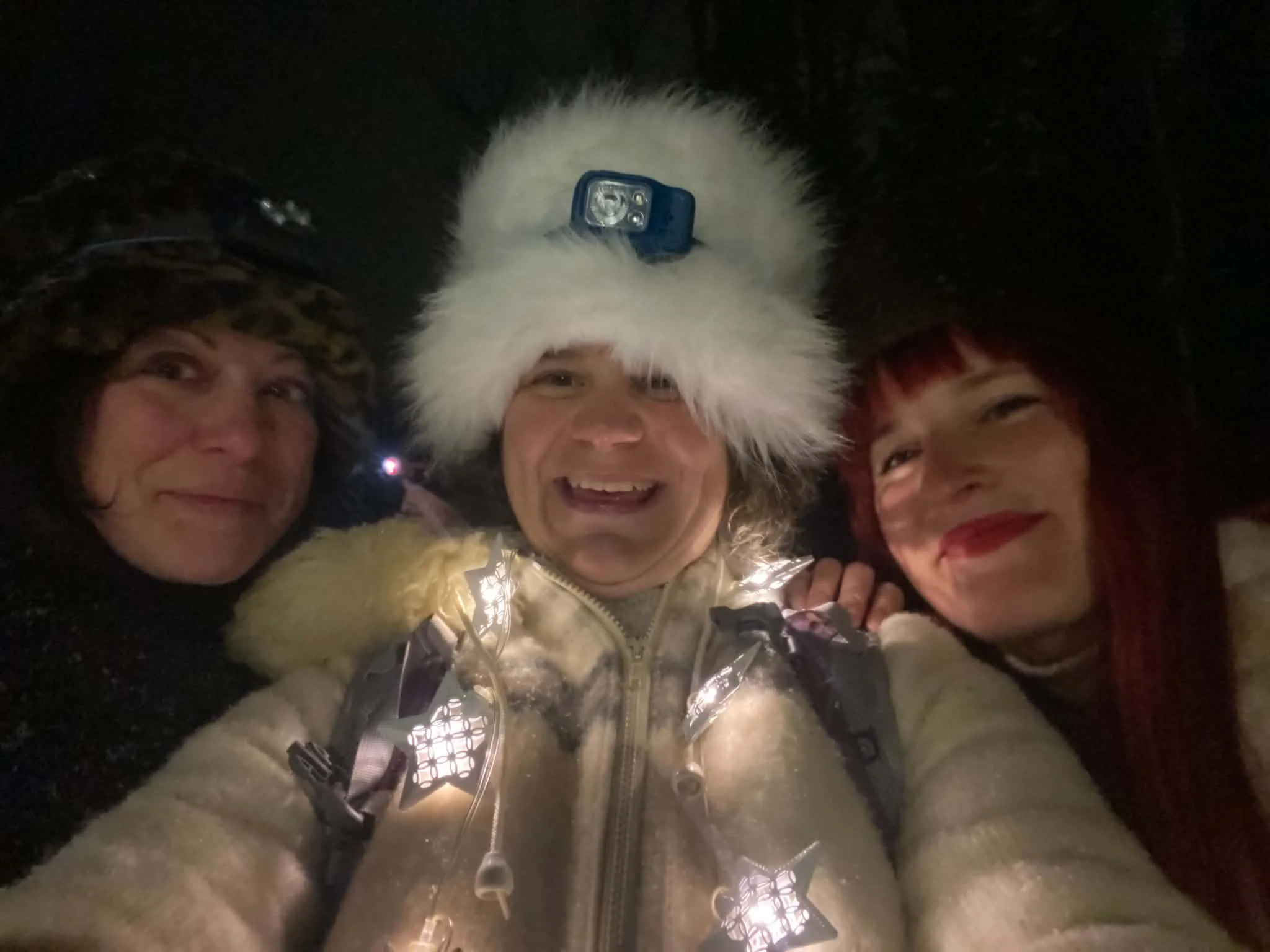 Three people outdoors at night, smiling, with one person wearing a fuzzy hat with a headlamp, surrounded by string lights.