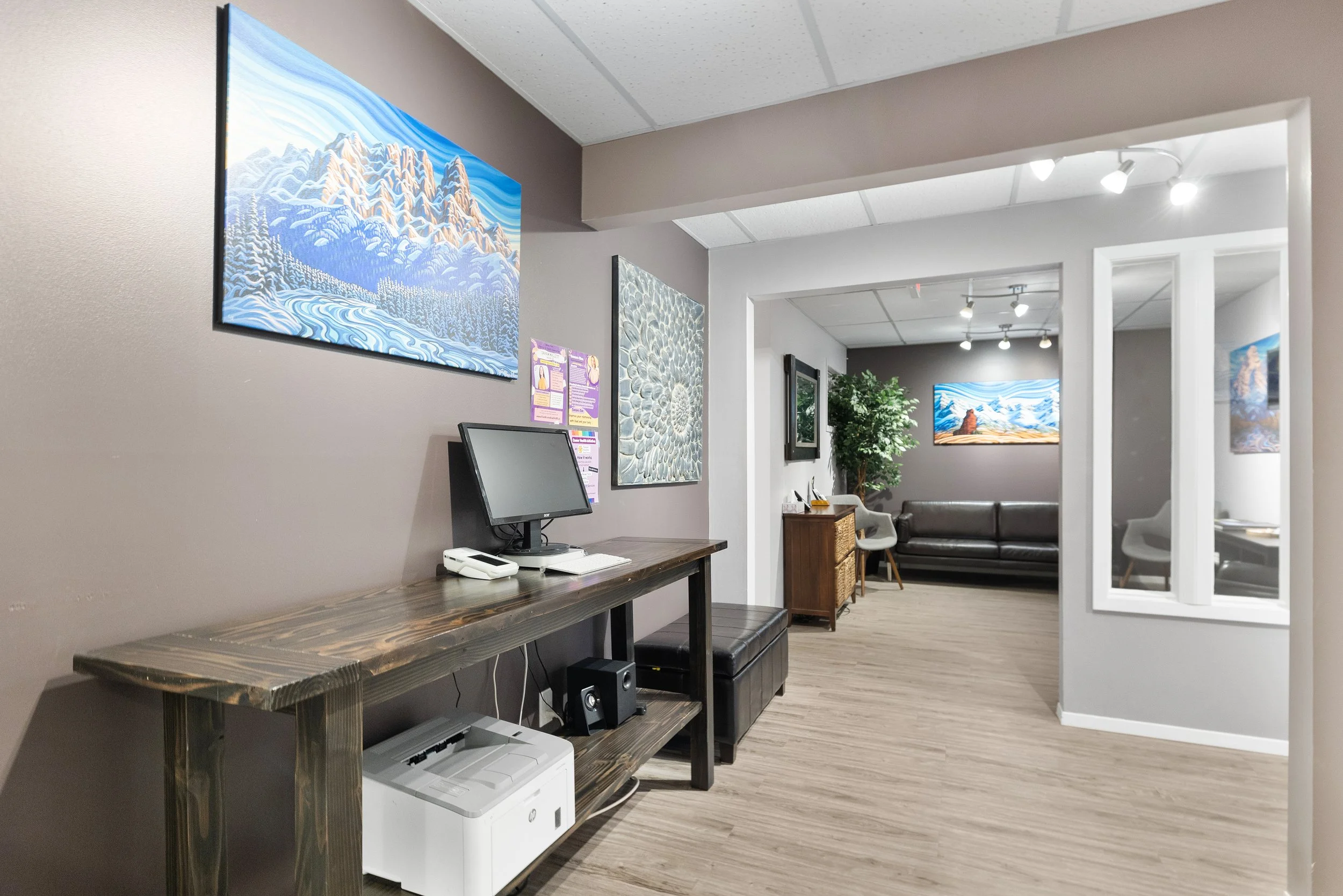A waiting room or hallway area with artwork on the walls, a small desk with a computer monitor and printer, and a sitting area with a black couch and chairs. There are track lights on the ceiling and a potted plant in the corner.