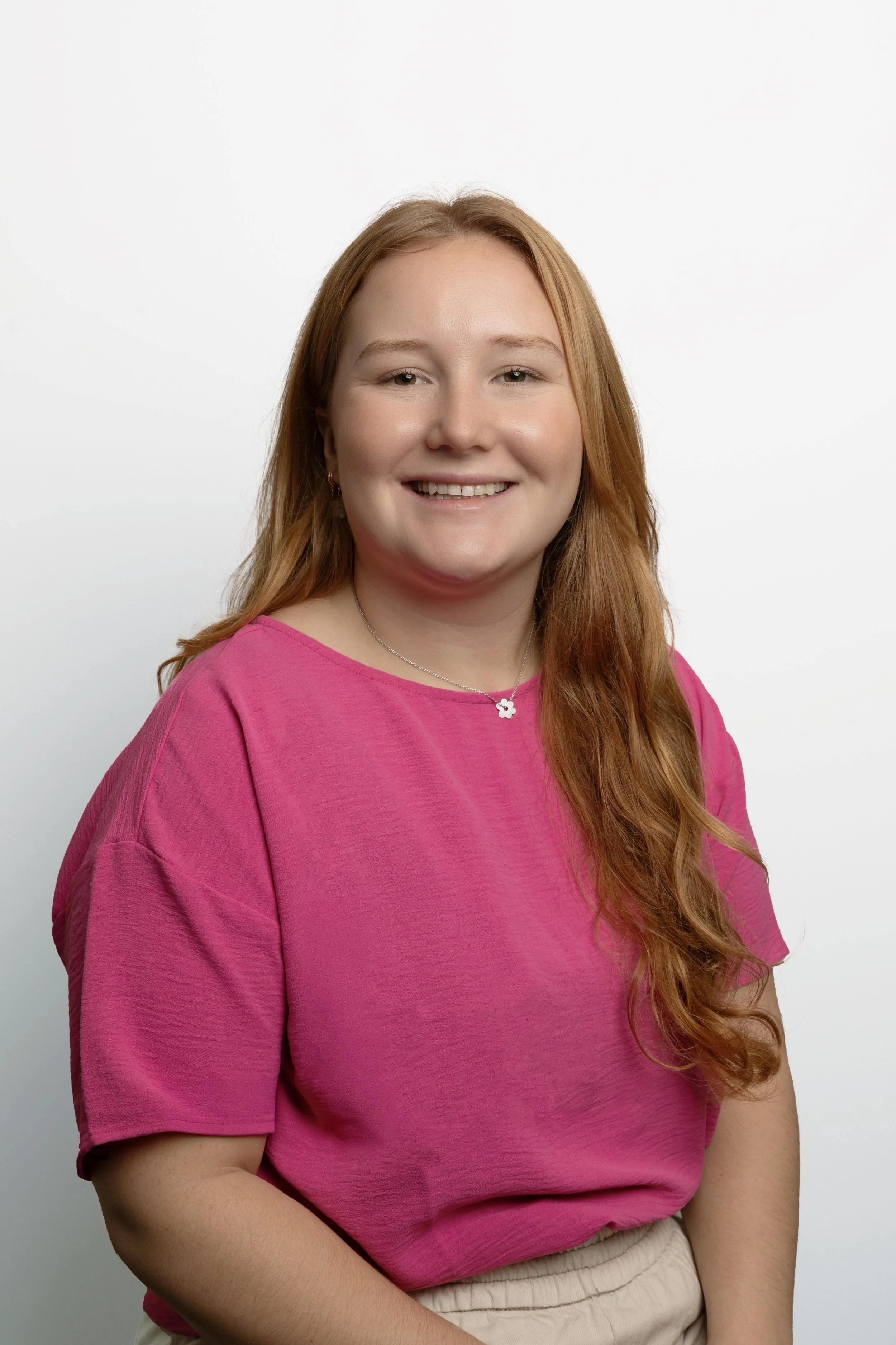 A young woman with long red hair and light skin, wearing a pink T-shirt and beige pants, smiling against a plain white background.