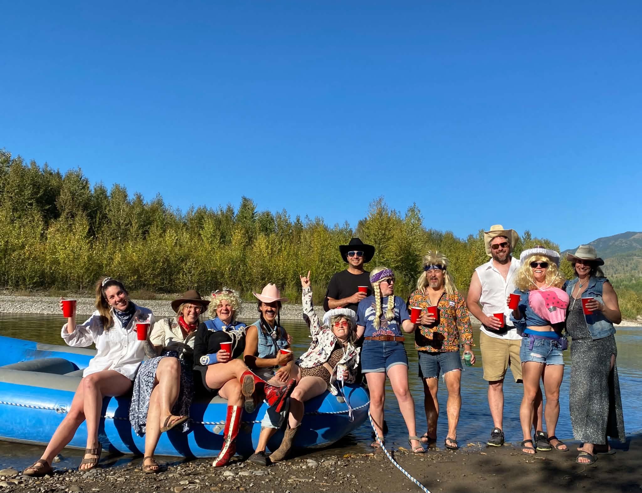 A group of people dressed in costume, smiling and holding red cups, gathered outdoors near a body of water with trees and mountains in the background on a sunny day.