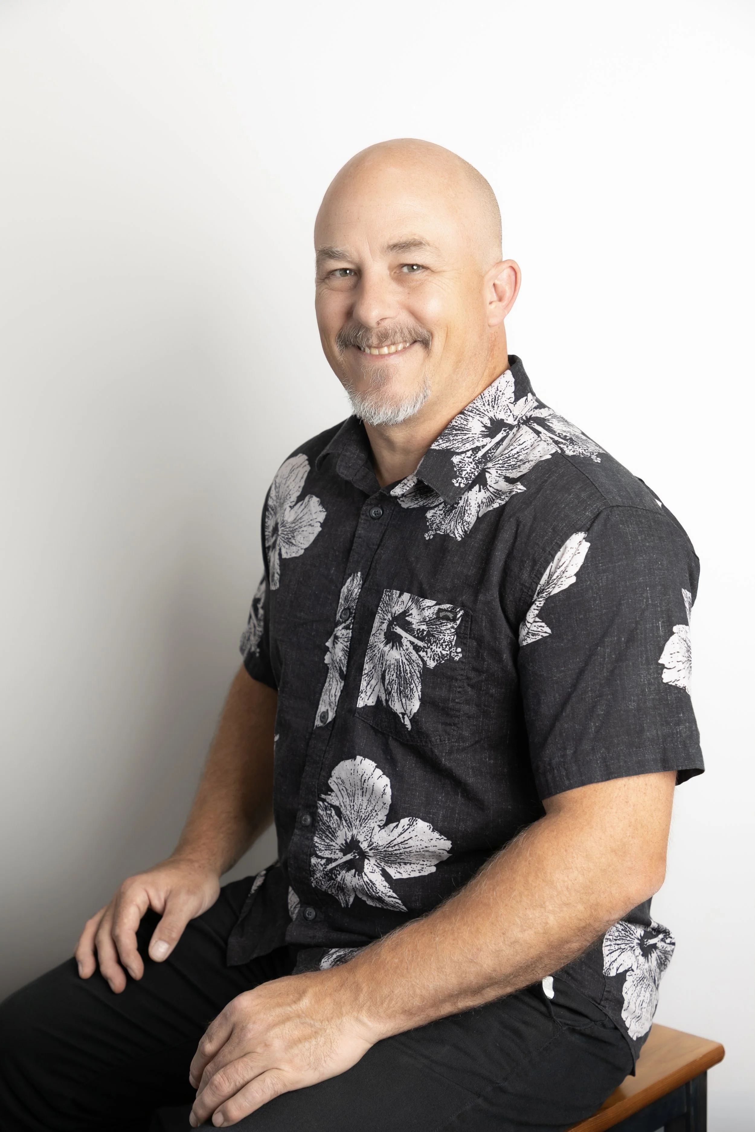 A smiling bald man with a goatee, wearing a black shirt with white floral pattern, sitting on a wooden bench against a plain white background.