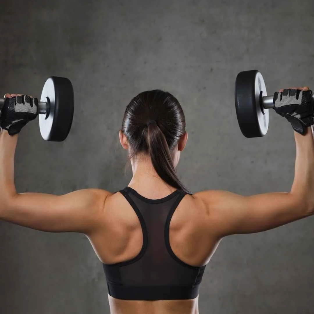 Back view of a woman with dark hair in a ponytail lifting dumbbells in a gym.
