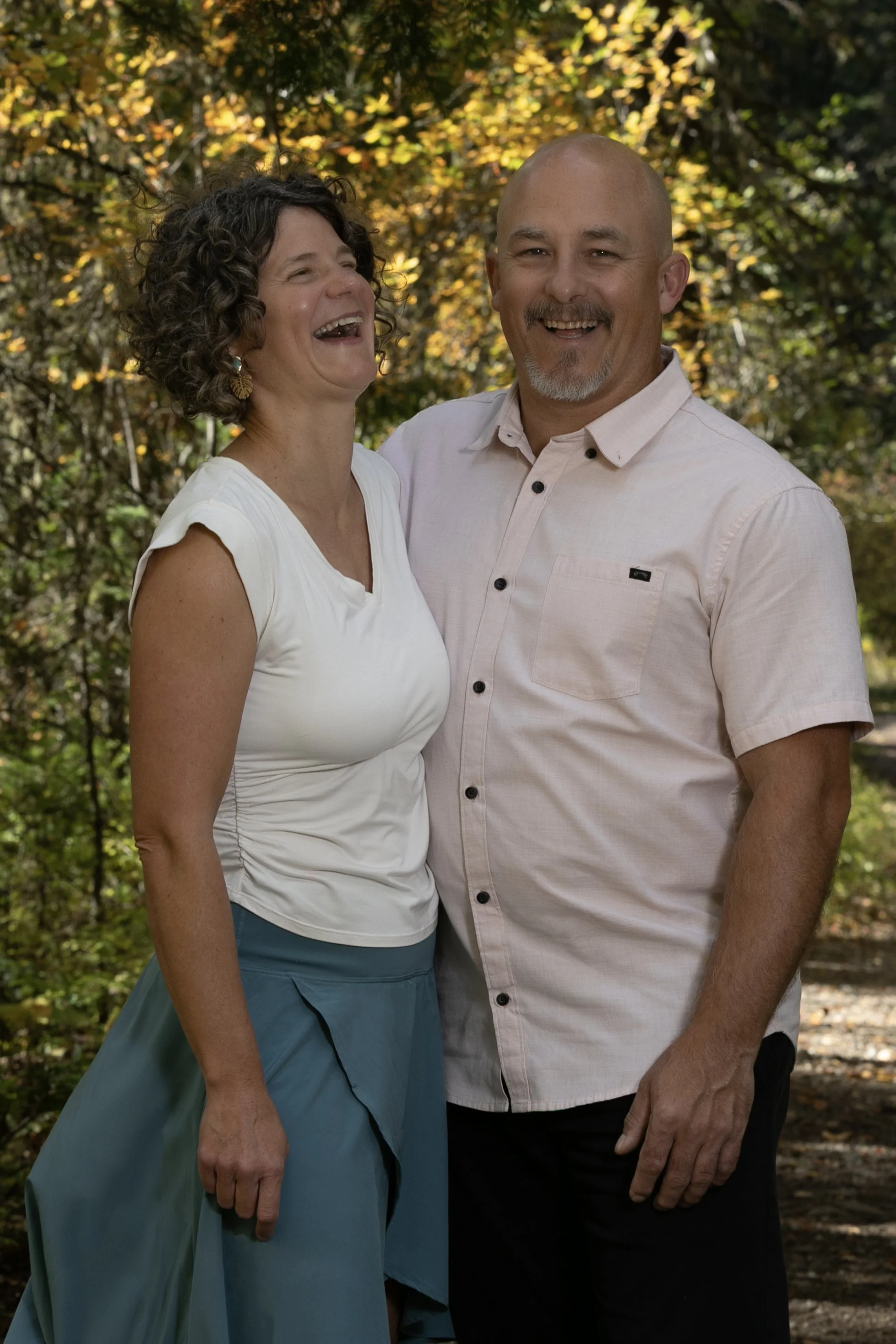 A smiling woman with curly dark hair and earrings, and a smiling man with a bald head and goatee, standing outdoors among fall foliage.