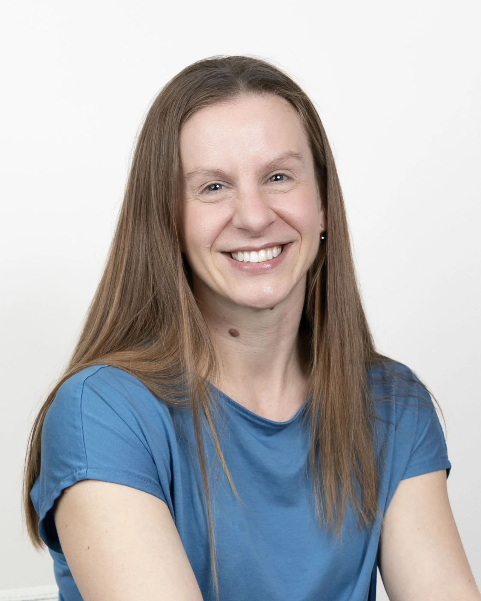 A woman with long reddish-brown hair smiling, wearing a blue t-shirt, against a plain white background.