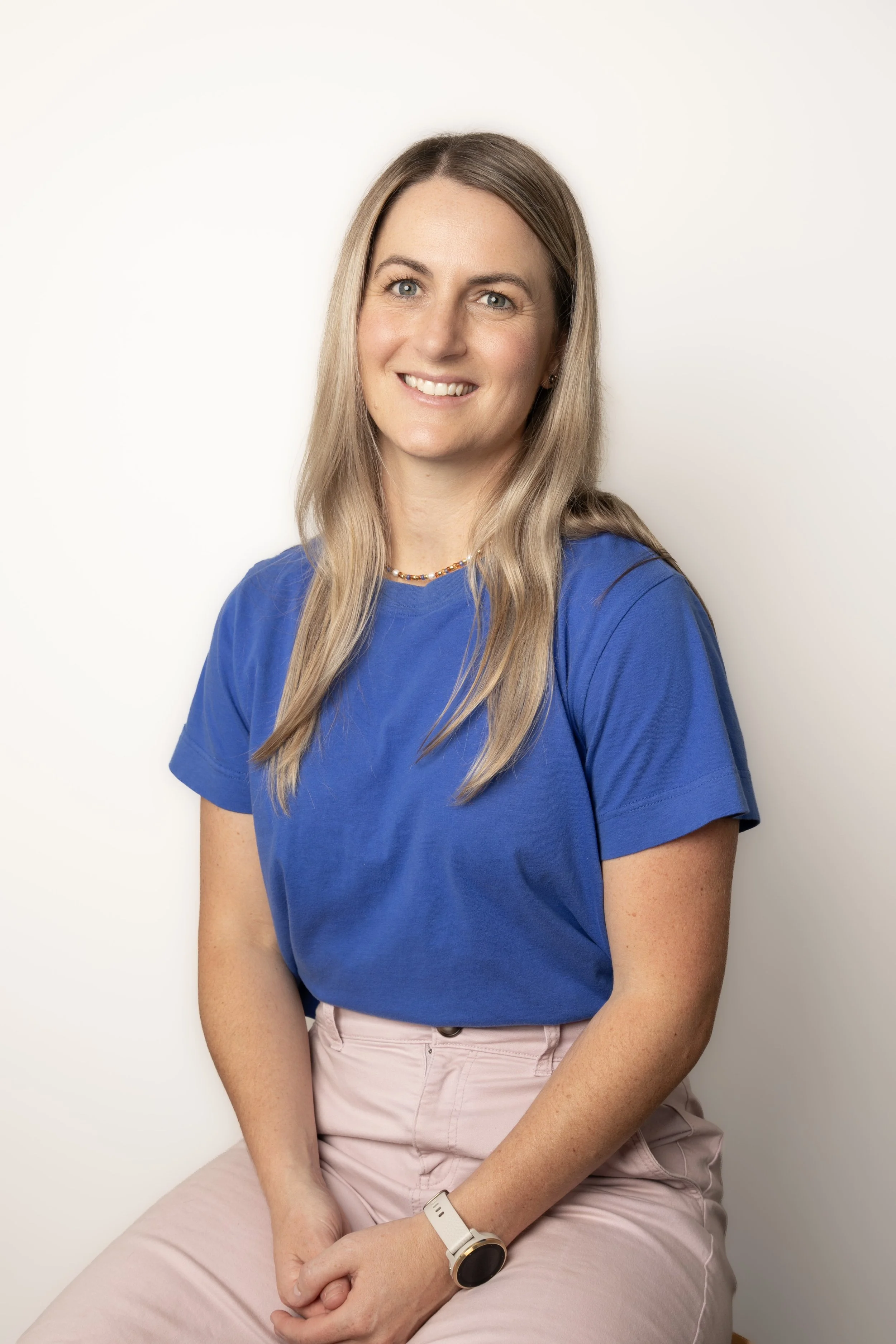 A woman with long blonde hair, blue eyes, wearing a blue t-shirt, pink pants, and a smartwatch, sitting against a plain white background.