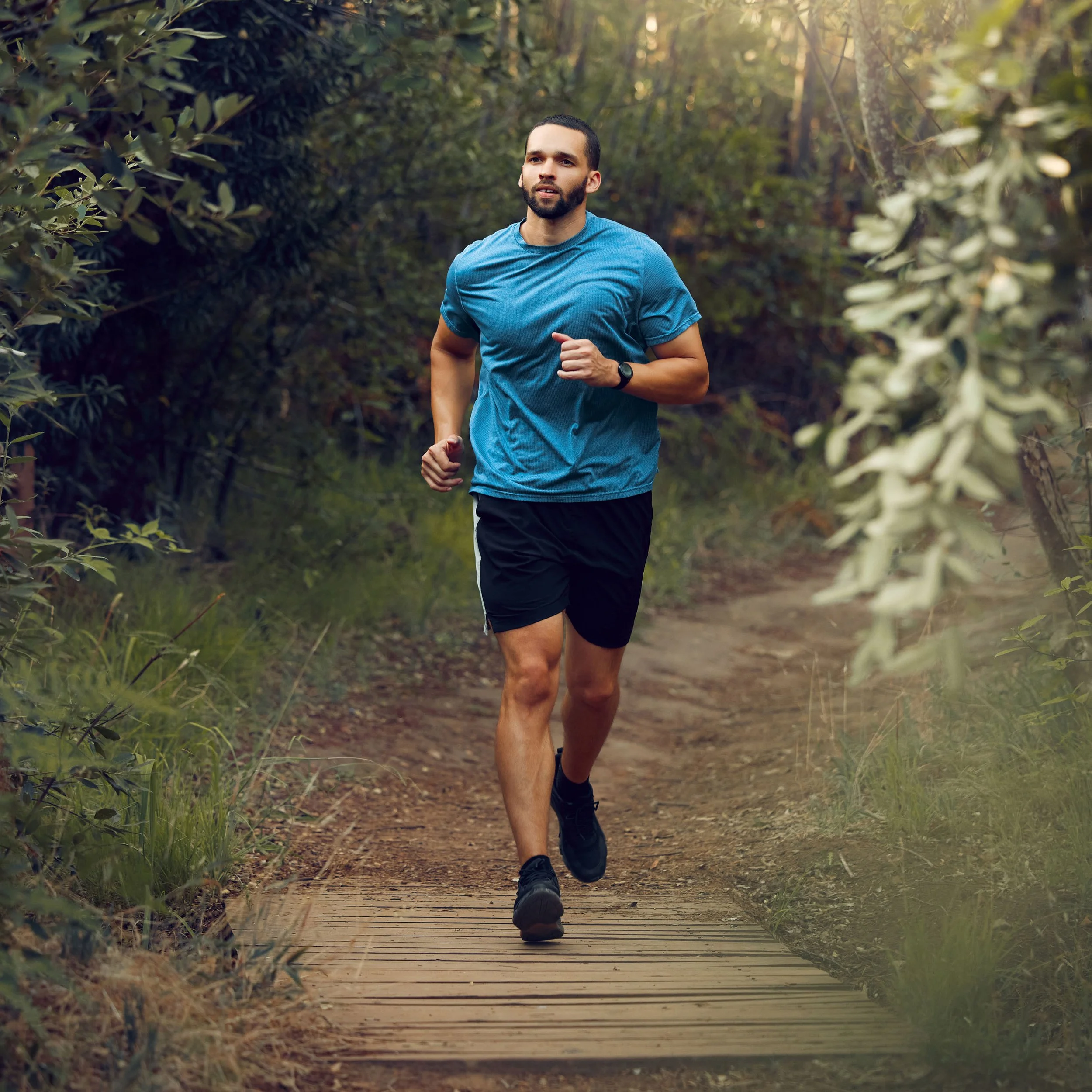 Man in a blue athletic shirt running on a dirt trail through a wooded area with greenery on both sides.