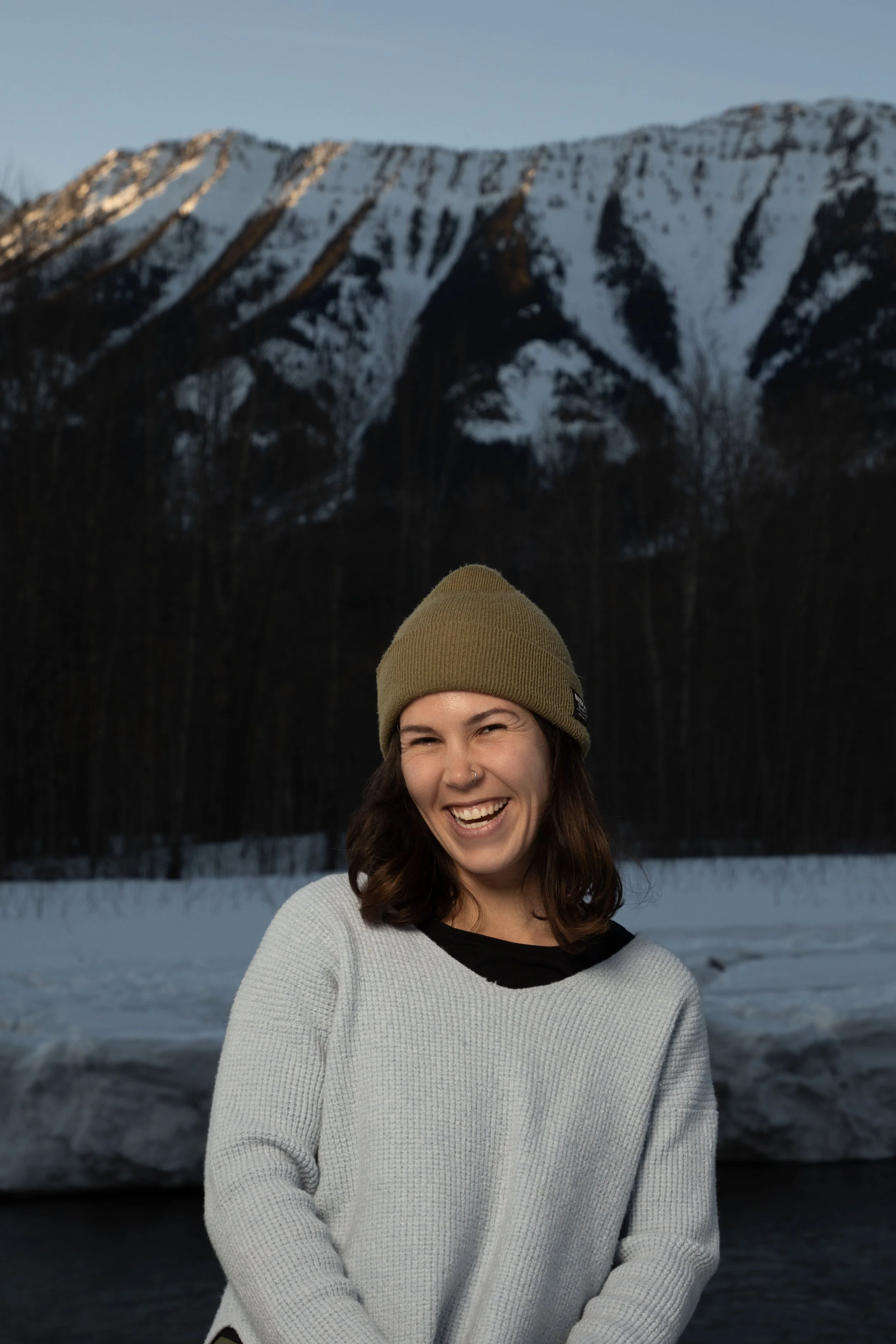 A young woman with brown hair smiling, wearing a green beanie and a white sweater, standing in front of a snowy landscape with mountains.