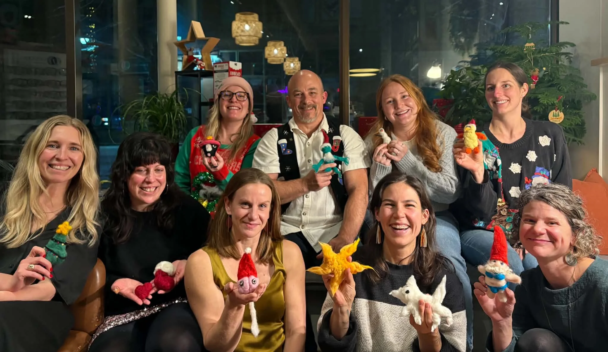 Group of people celebrating Christmas indoors, holding handmade Christmas ornaments and plush decorations, smiling and wearing festive sweaters and hats.