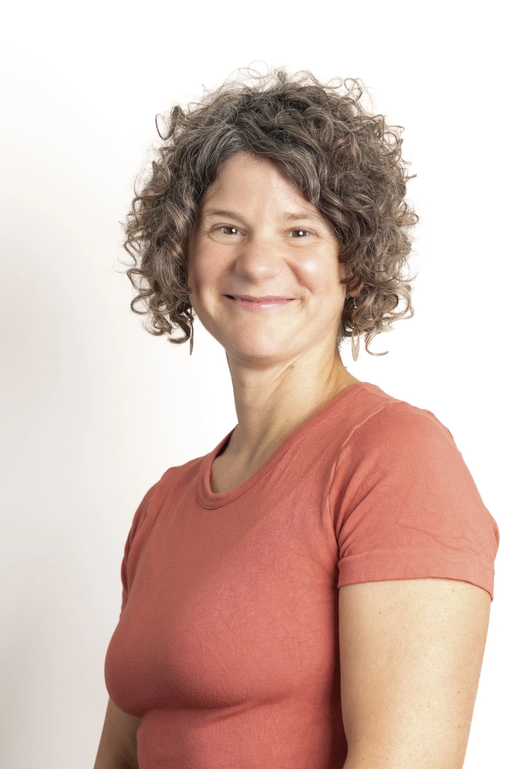 A woman with short curly hair smiling, wearing a rust-colored shirt, against a plain white background.