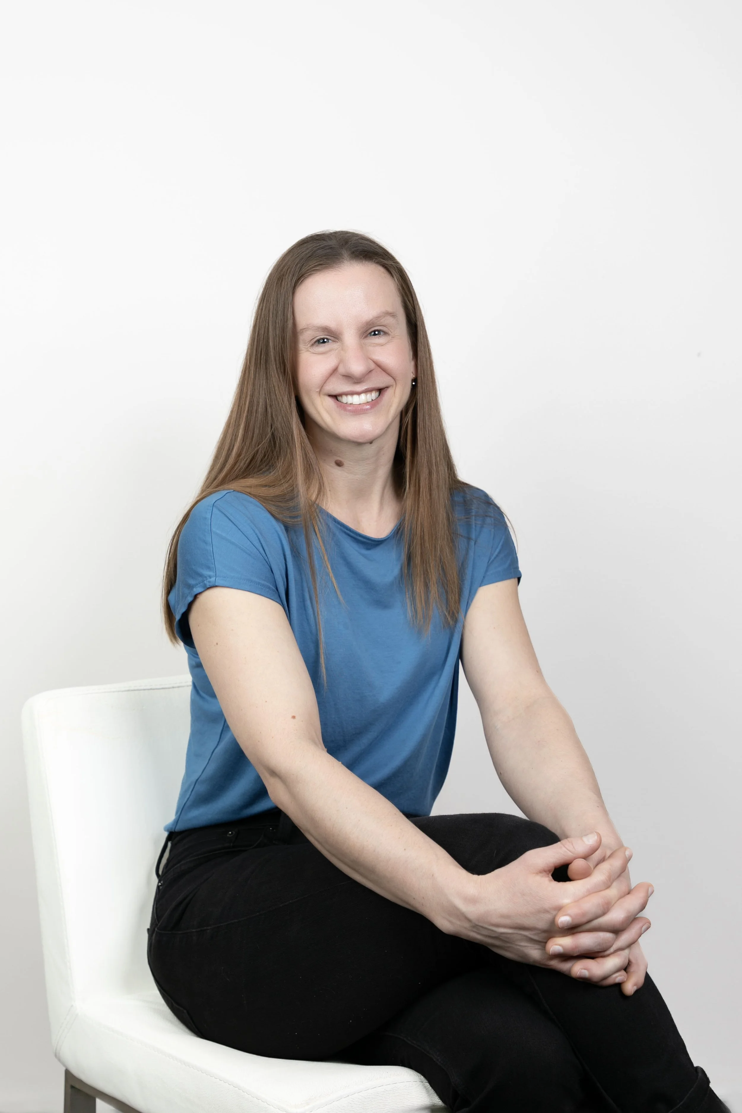 A woman with long brown hair, wearing a blue shirt and black pants, sitting on a white chair against a plain white background.