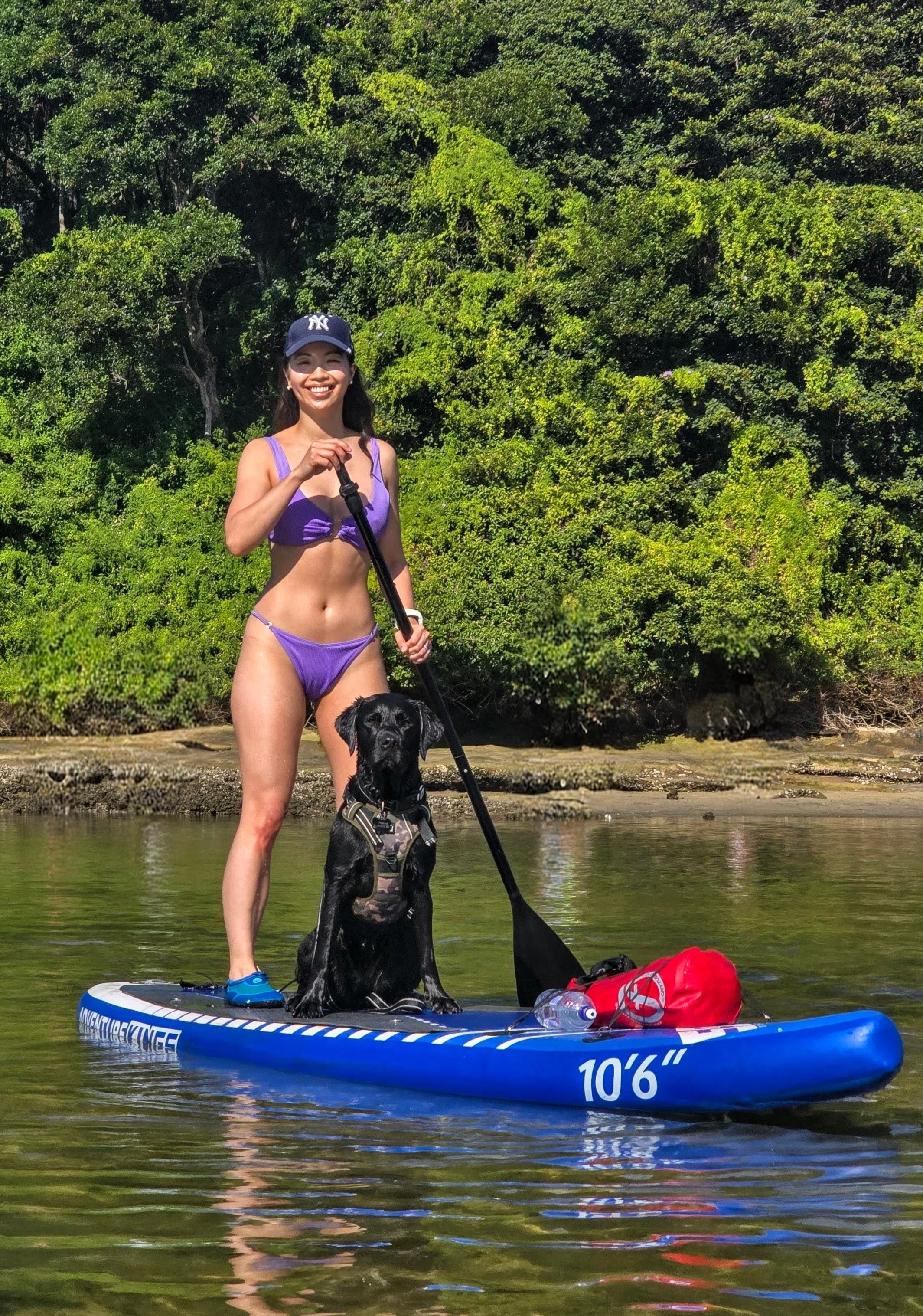 A woman in a purple bikini and a New York Yankees cap stands on an inflatable paddleboard with a black dog in a harness. She holds a paddle and smiles at the camera with a background of lush green trees and water.