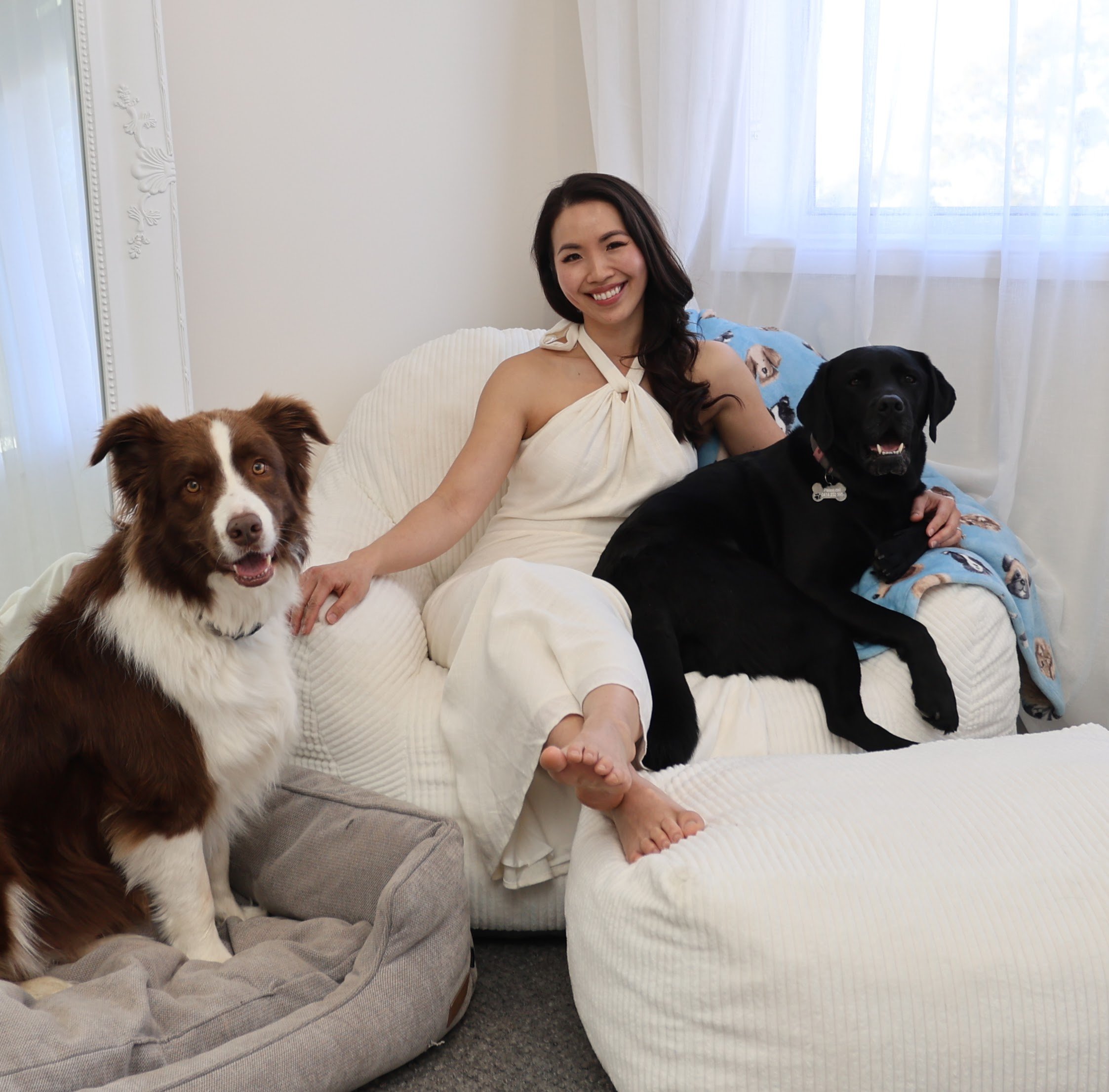 A woman smiling while sitting on a bed with her hands resting on her lap, accompanied by three dogs; a brown and white dog with pointy ears and a happy expression, a black dog with a collar, and another brown dog partially visible, in a well-lit bedroom with white walls, a window with curtains, and a white bedspread.