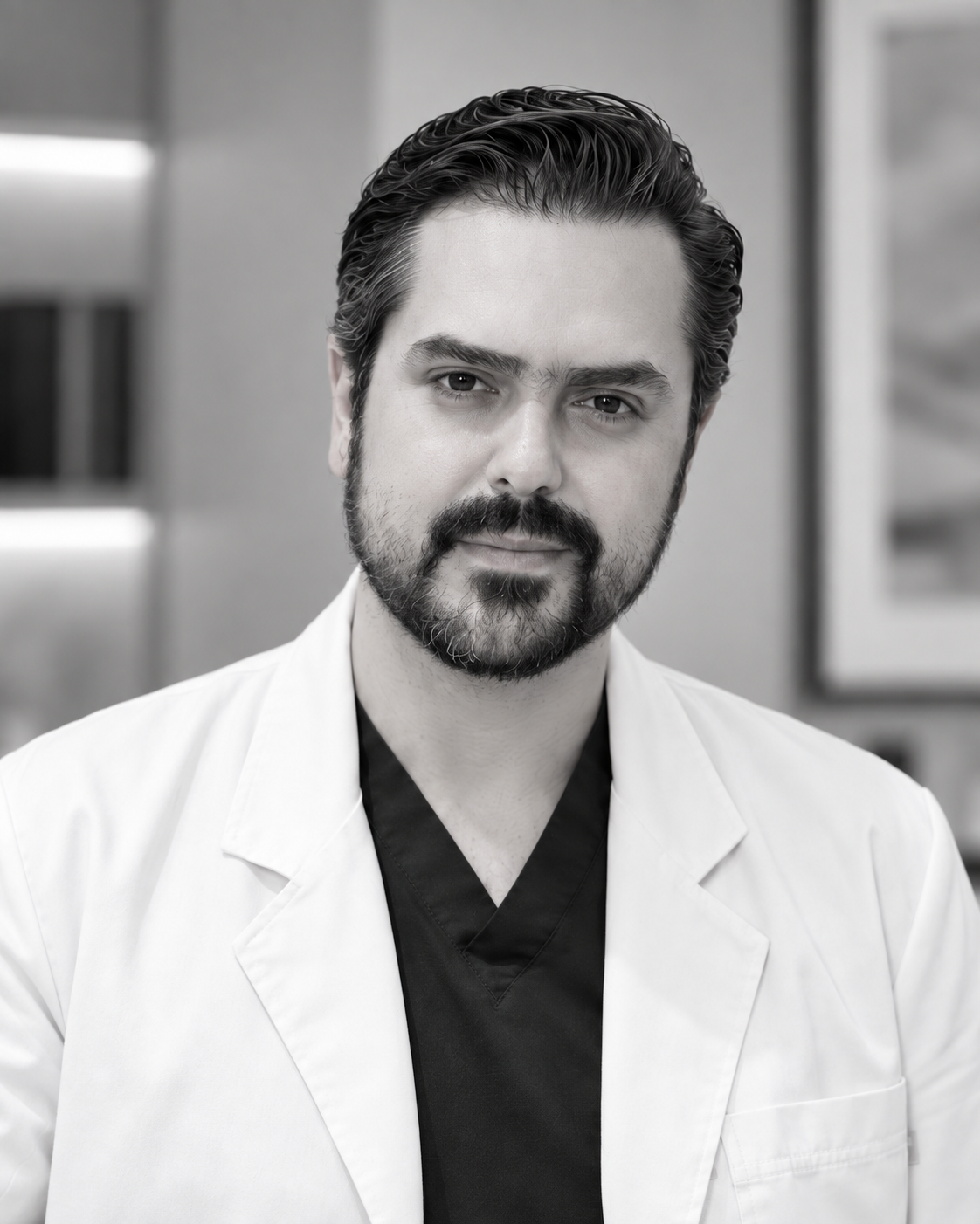 A man with dark hair, a beard, and mustache wearing a white lab coat and black shirt, standing in a medical or laboratory setting.