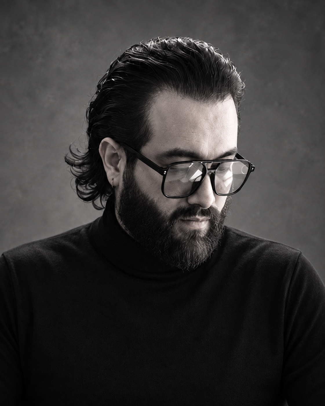A black and white portrait of a man with dark hair styled back, a beard, wearing large glasses and a black turtleneck, looking downward with a serious expression.