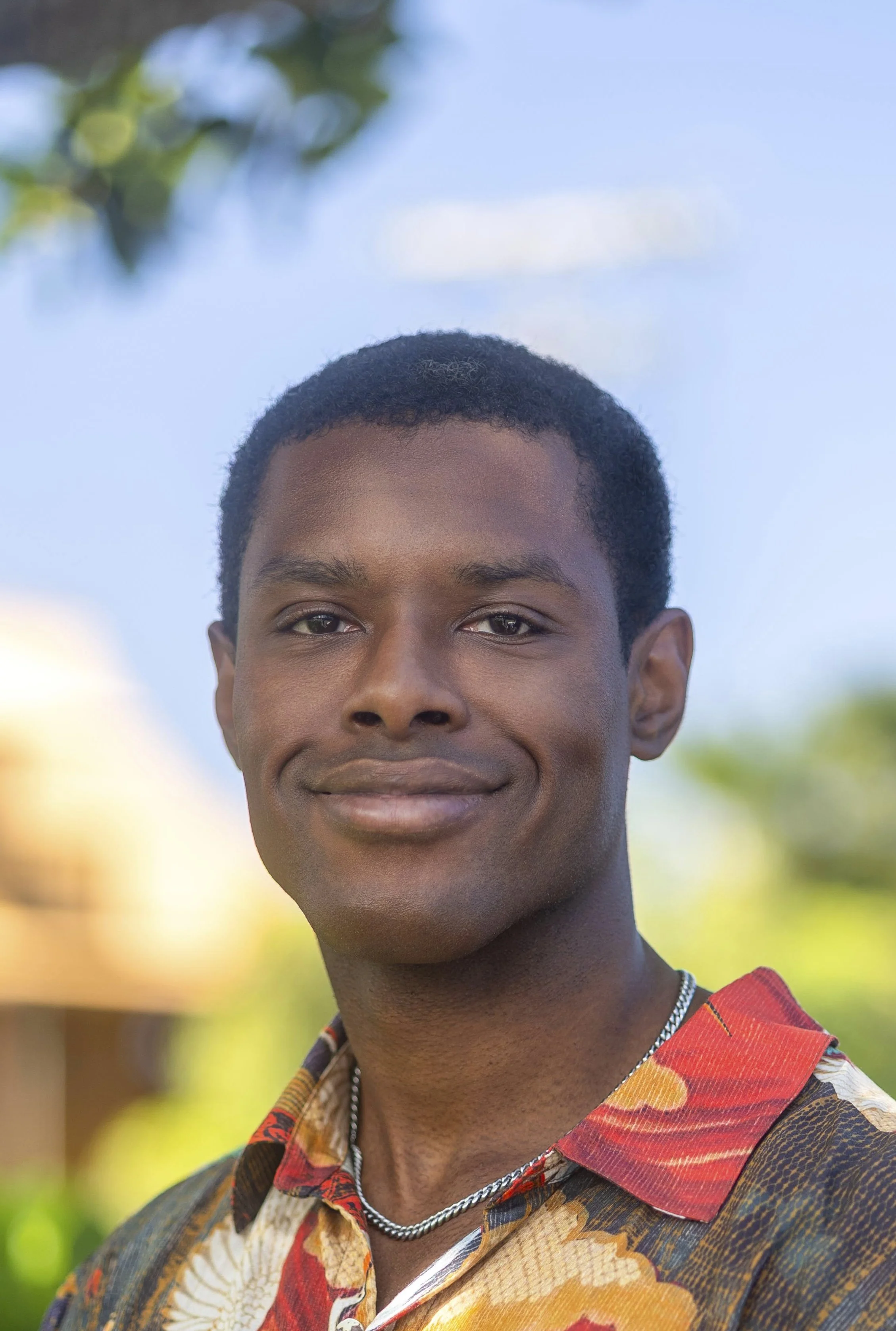 Close-up portrait of a young man with short black hair, smiling slightly, wearing a colorful patterned shirt, a silver chain necklace, and standing outdoors with a blue sky and blurred greenery in the background.