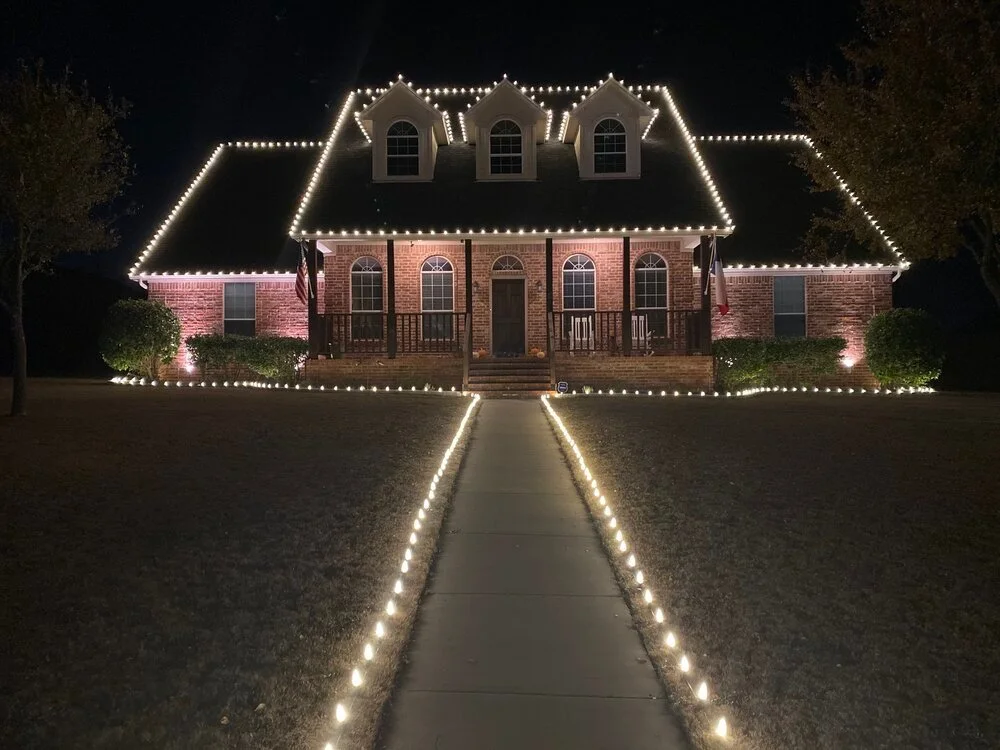 A large brick house decorated with white string lights outlining the roof, windows, and walkway at night.