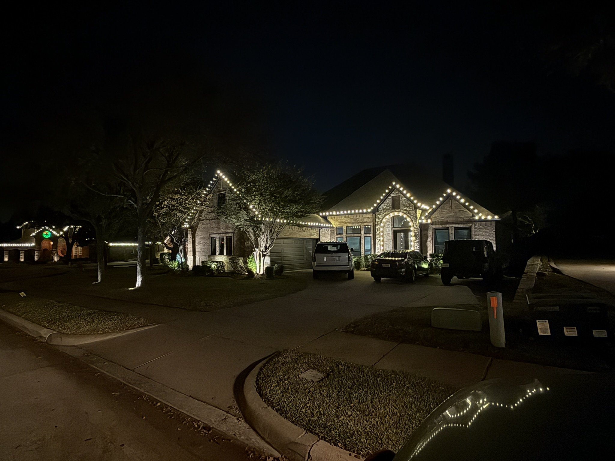 Nighttime scene of a residential neighborhood with houses decorated with Christmas lights. The house in the center has string lights outlining the roof and around the entrance. Several vehicles are parked in the driveway and street. Trees and a well-maintained lawn are visible.
