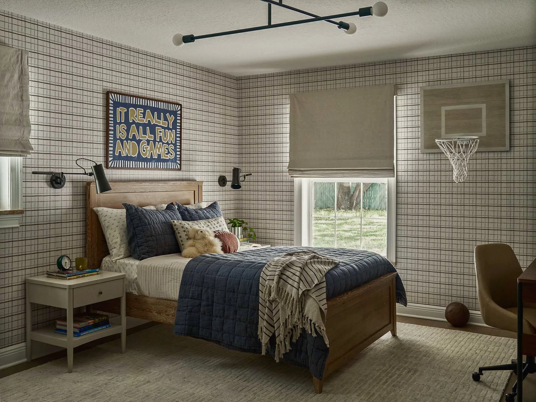 A bedroom with a checkered wall, a wooden bed with pillows and a blanket, a white nightstand with books, a window with blinds, a ceiling light fixture, and a small basketball hoop on the wall.