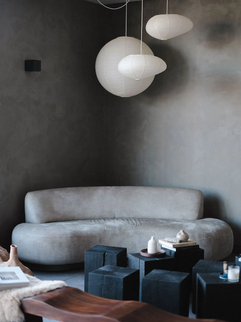 A modern living room featuring a beige curved sofa, black geometric coffee tables with candles and books, gray walls, and white paper lantern pendant lights hanging from the ceiling.
