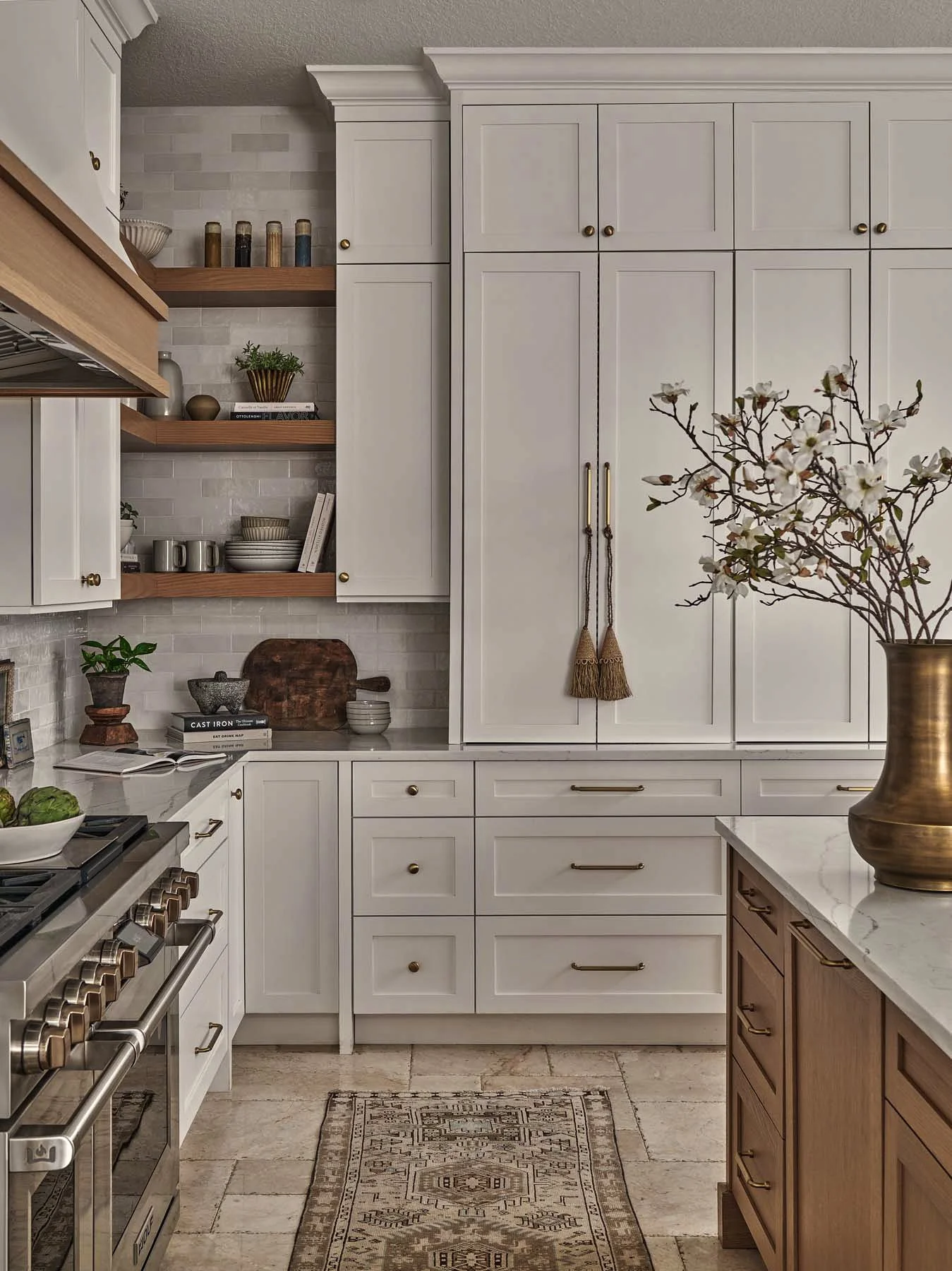 A kitchen with white cabinets, open wood shelves with spices, dishes, and decor, a marble countertop, a gold vase with flowering branches, and a patterned rug on beige tile floor.