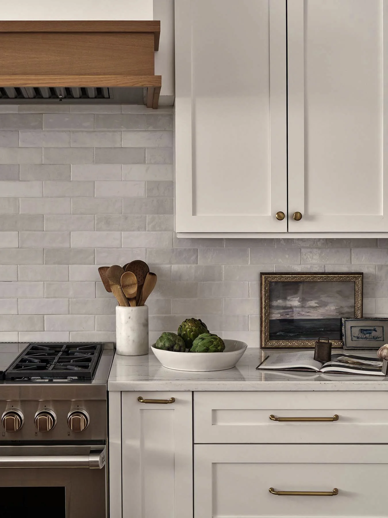 Kitchen countertop with a white marble surface, a ceramic utensil holder with wooden spoons and a spatula, a bowl of artichokes, an open book, framed artwork, and part of a stove with stainless steel oven.