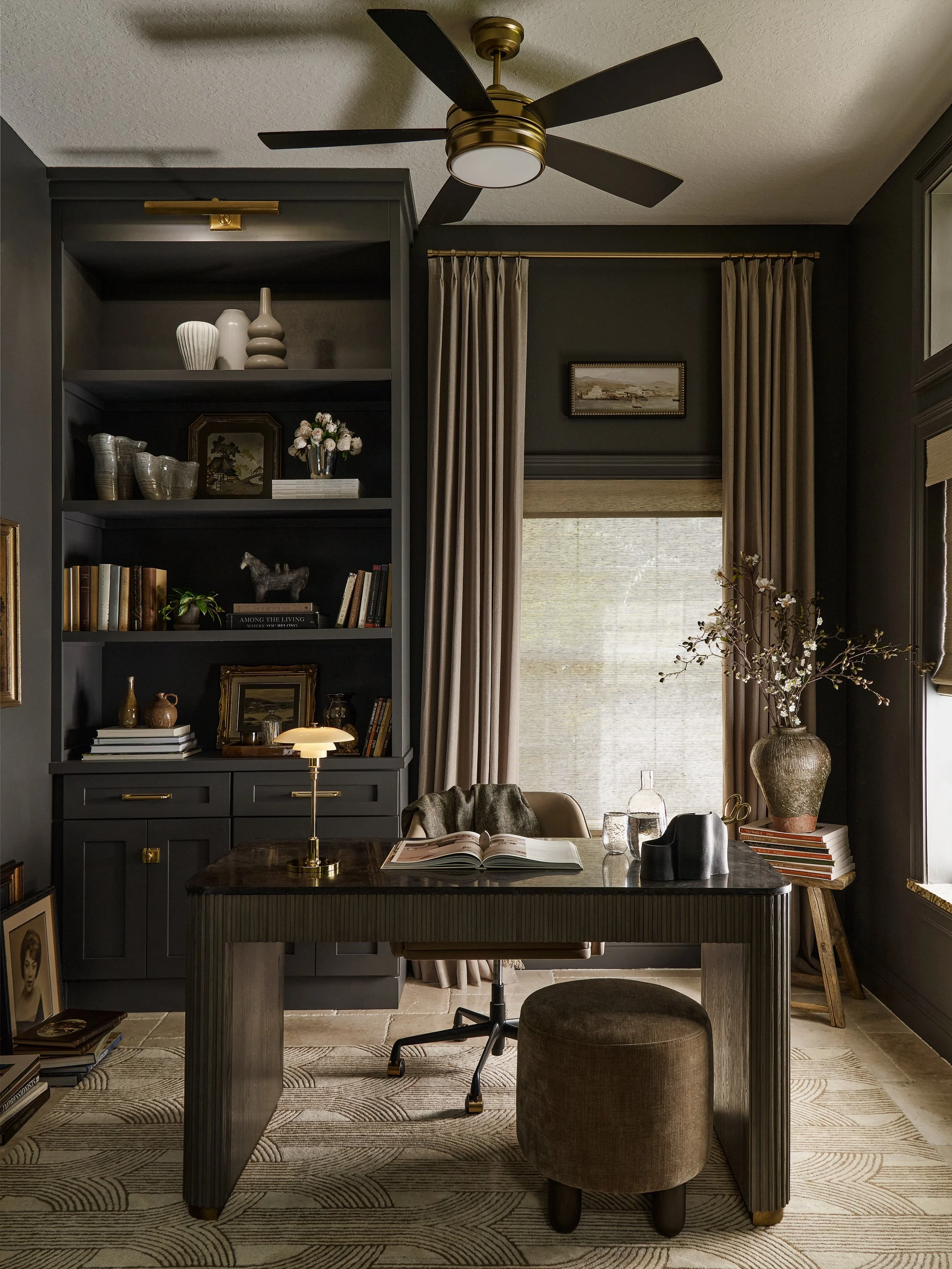 A home office with a dark gray built-in bookshelf, a desk with a lit table lamp, open book, and decorative objects, beige curtains, a large window, and a potted plant in a textured vase.
