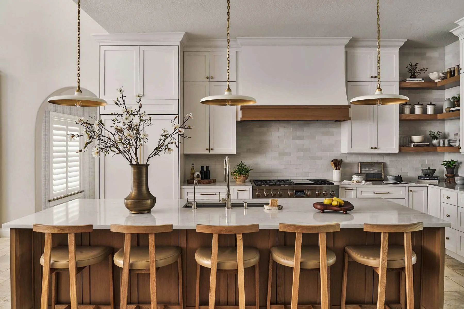 A modern kitchen with a white marble island, wooden bar stools, white cabinetry, open shelves with dishes and decor, a stainless steel stove, and a vase with branches and flowers on the island.