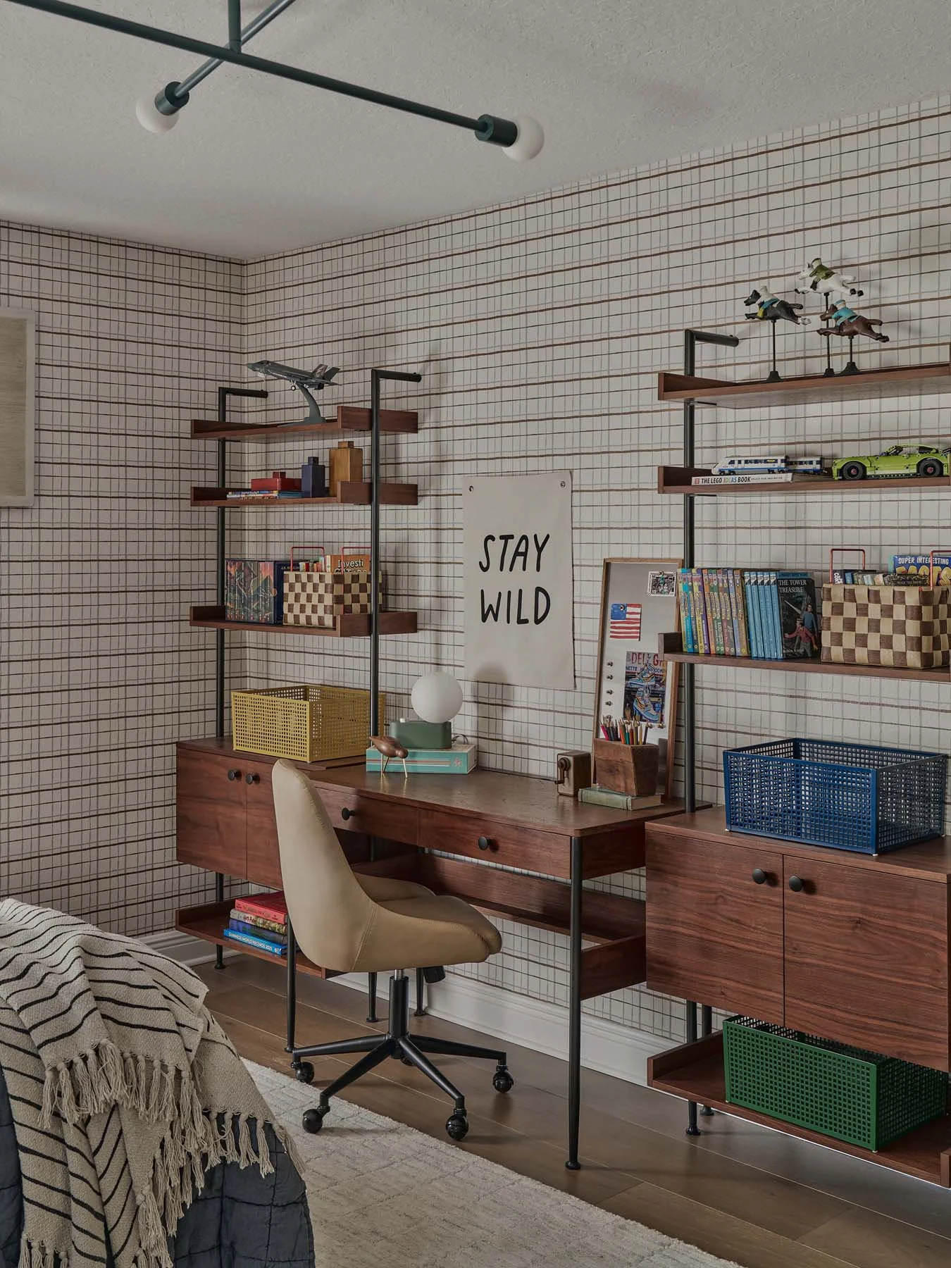 A home office with grid-patterned wallpaper, wooden desk, beige swivel chair, multiple shelves with books and decorative items, and a sign that says "STAY WILD" on the wall.
