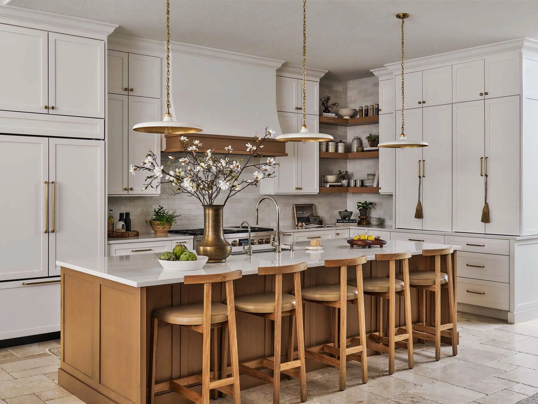 Modern kitchen with white cabinets, a large kitchen island with wooden chairs, a brass vase with white flowers, and open wooden shelves with various kitchenware.