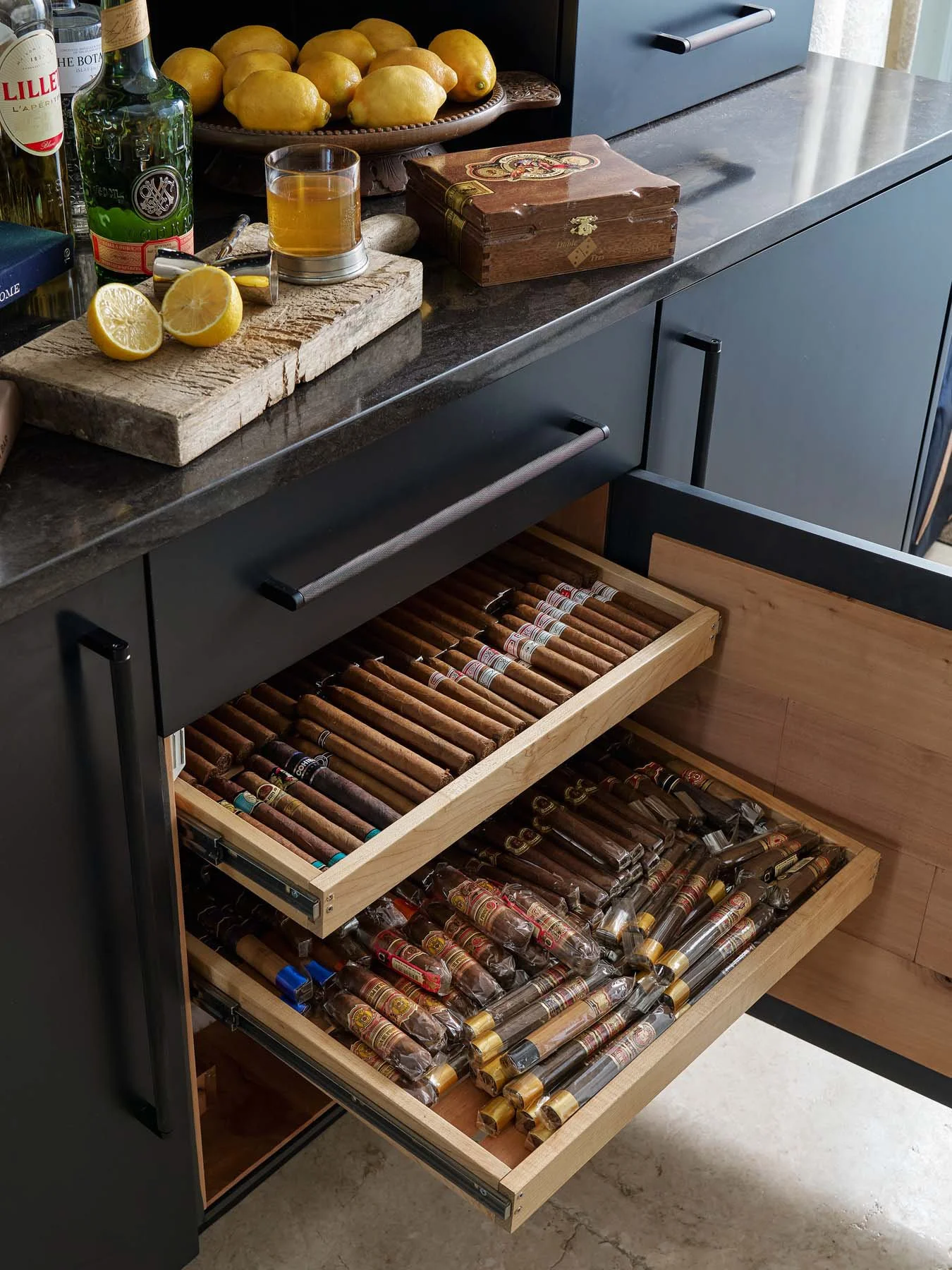 Kitchen drawer filled with cigars, found below a countertop with lemons, bottles of alcohol, and a glass of liquor on a cutting board.