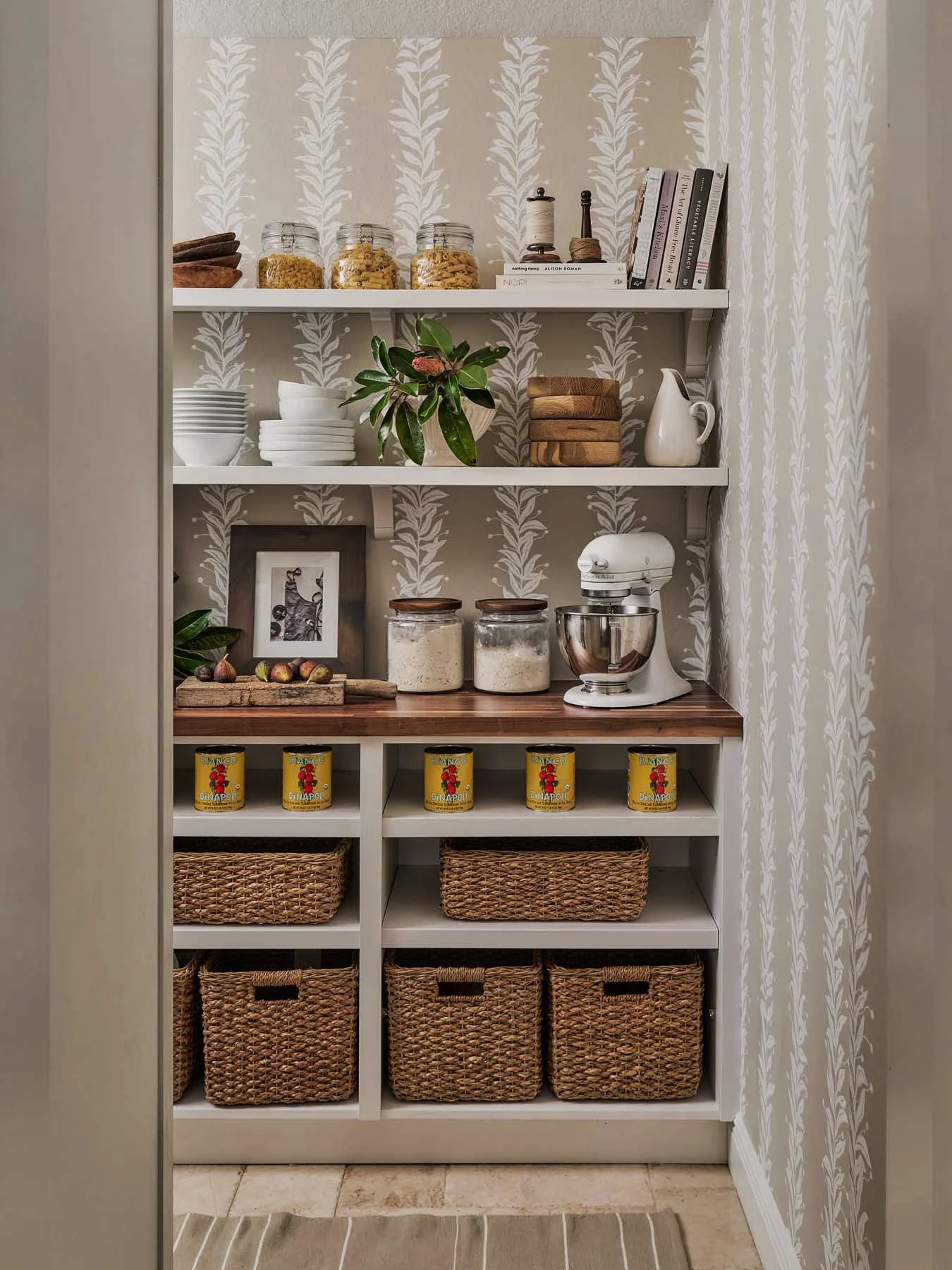 A white pantry with four shelves filled with jars, bowls, books, a plant, a mixer, canned goods, and woven baskets. The top shelf has jars of pasta and dry goods, a small book and decorative items. The second shelf has white bowls, a plant, wooden bo