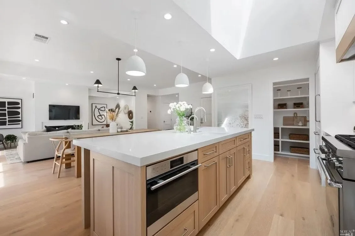 Modern kitchen with light wood cabinets and white countertop, open layout with living area, minimalist decor, pendant lighting, and stainless steel appliances.