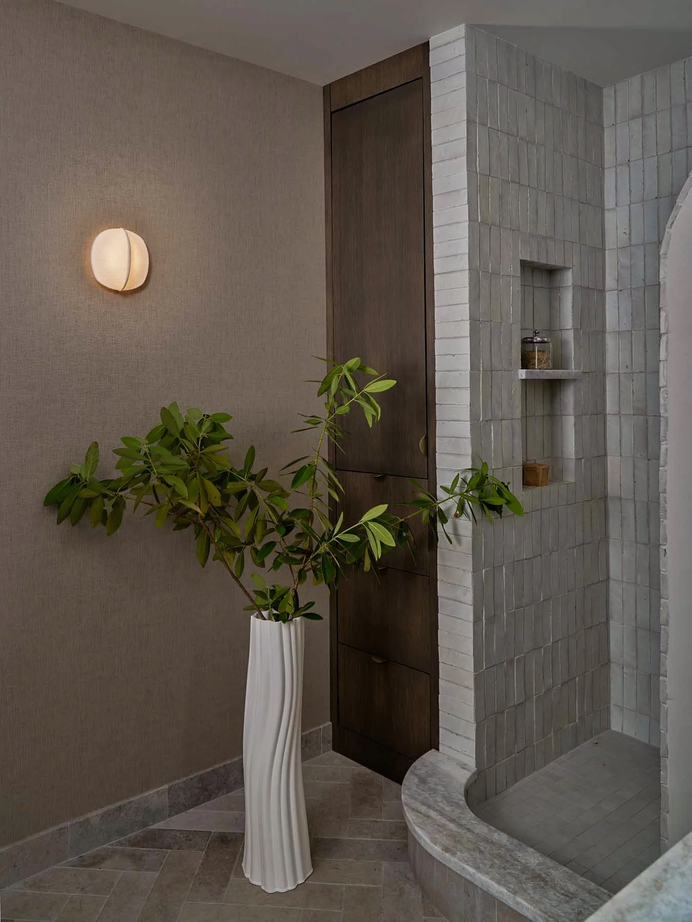 A modern interior space with a textured beige wall, a white lounge wall sconce, a tall white orchid plant in a white vase, and a corner displaying a brick and wood structure.