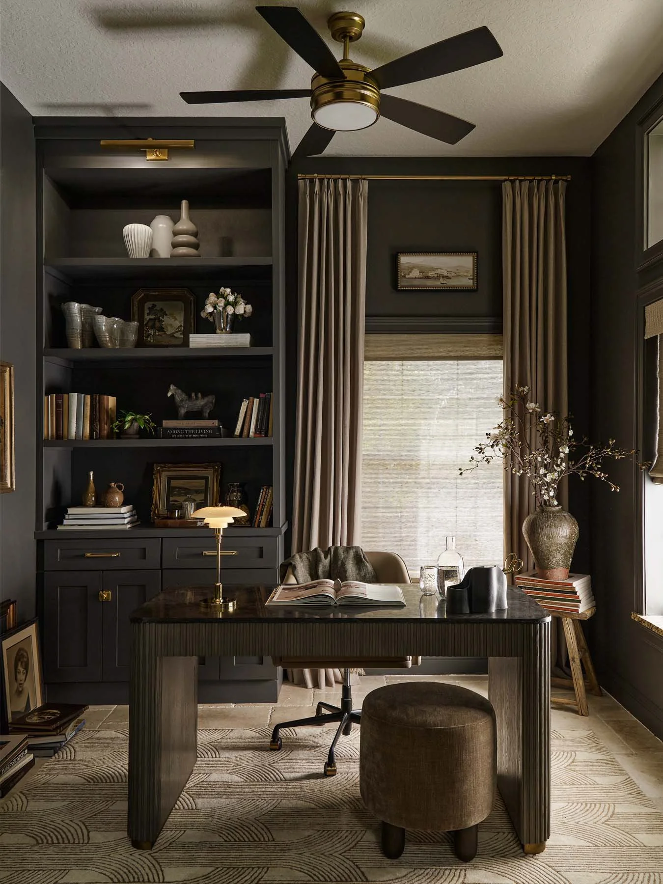 A home office or study room with dark-colored walls and a ceiling fan. There is a large window with beige curtains, a bookshelf filled with books and decorative items, and a desk with an open book, glassware, and a black decor piece. A small stool an